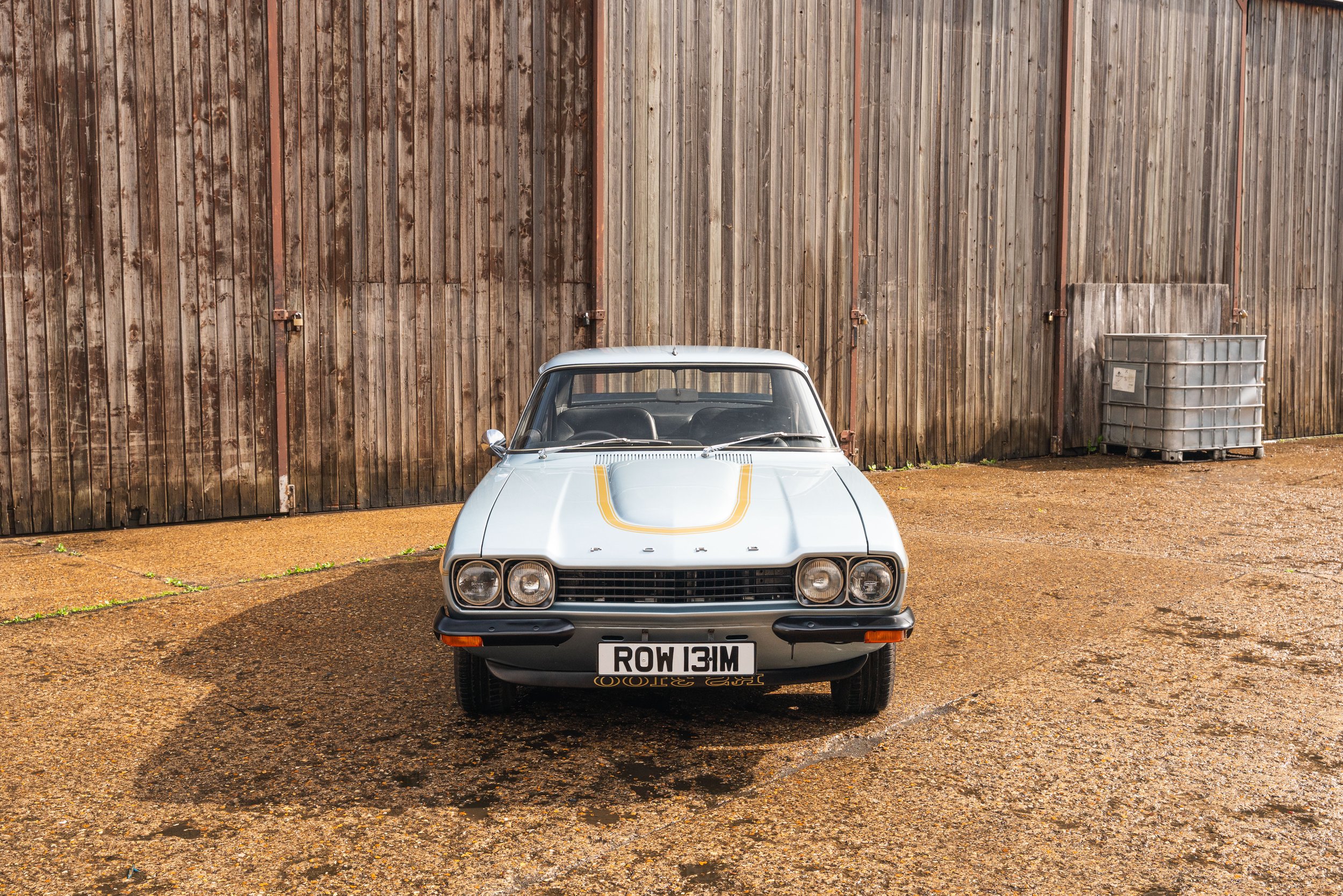 A vintage white coupe car with a yellow racing stripe on the hood, parked in front of a wooden fence, casting a shadow on the gravel ground.