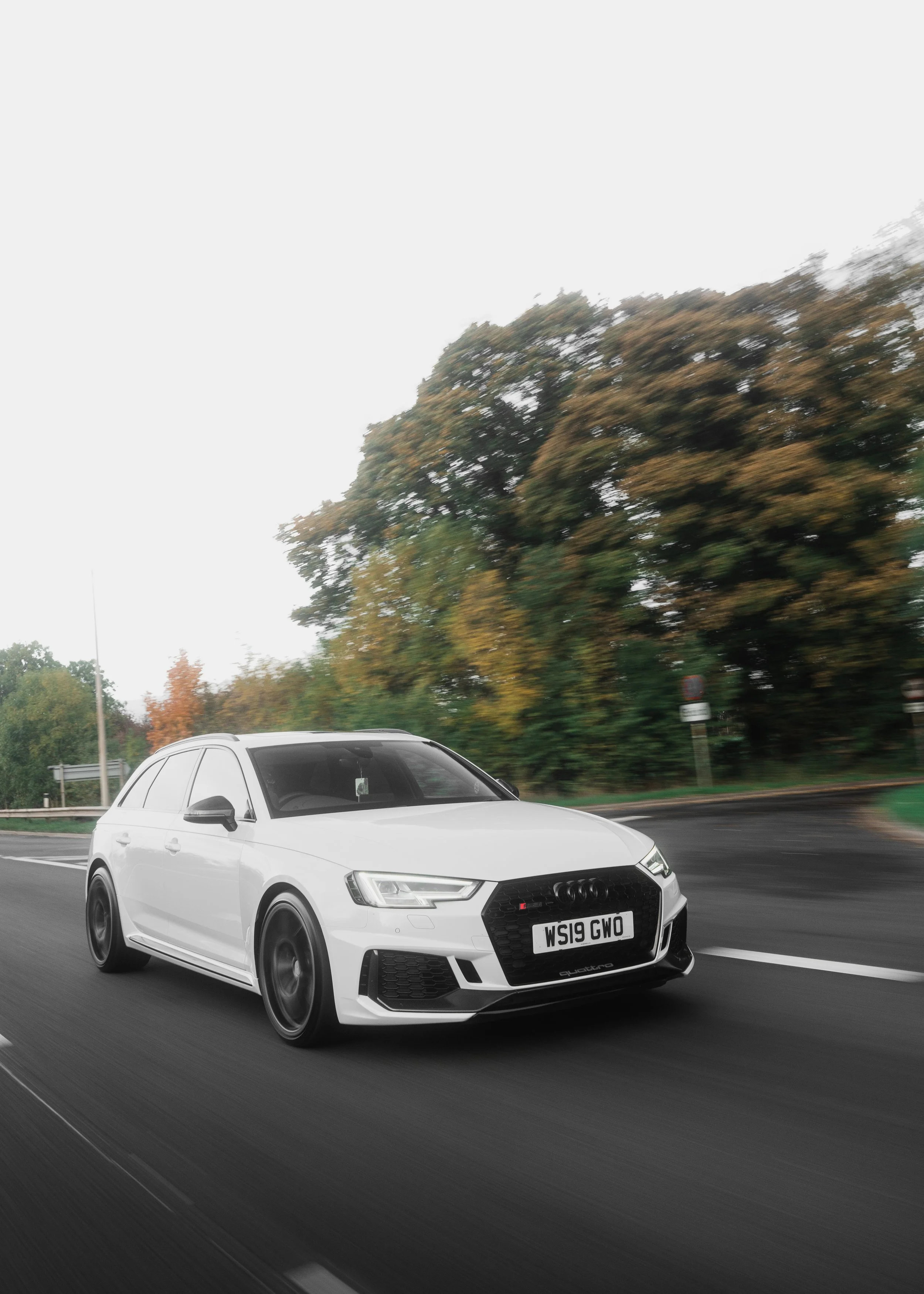 A white Audi RS4 Avant driving on a highway with blurred green trees and fall foliage in the background.