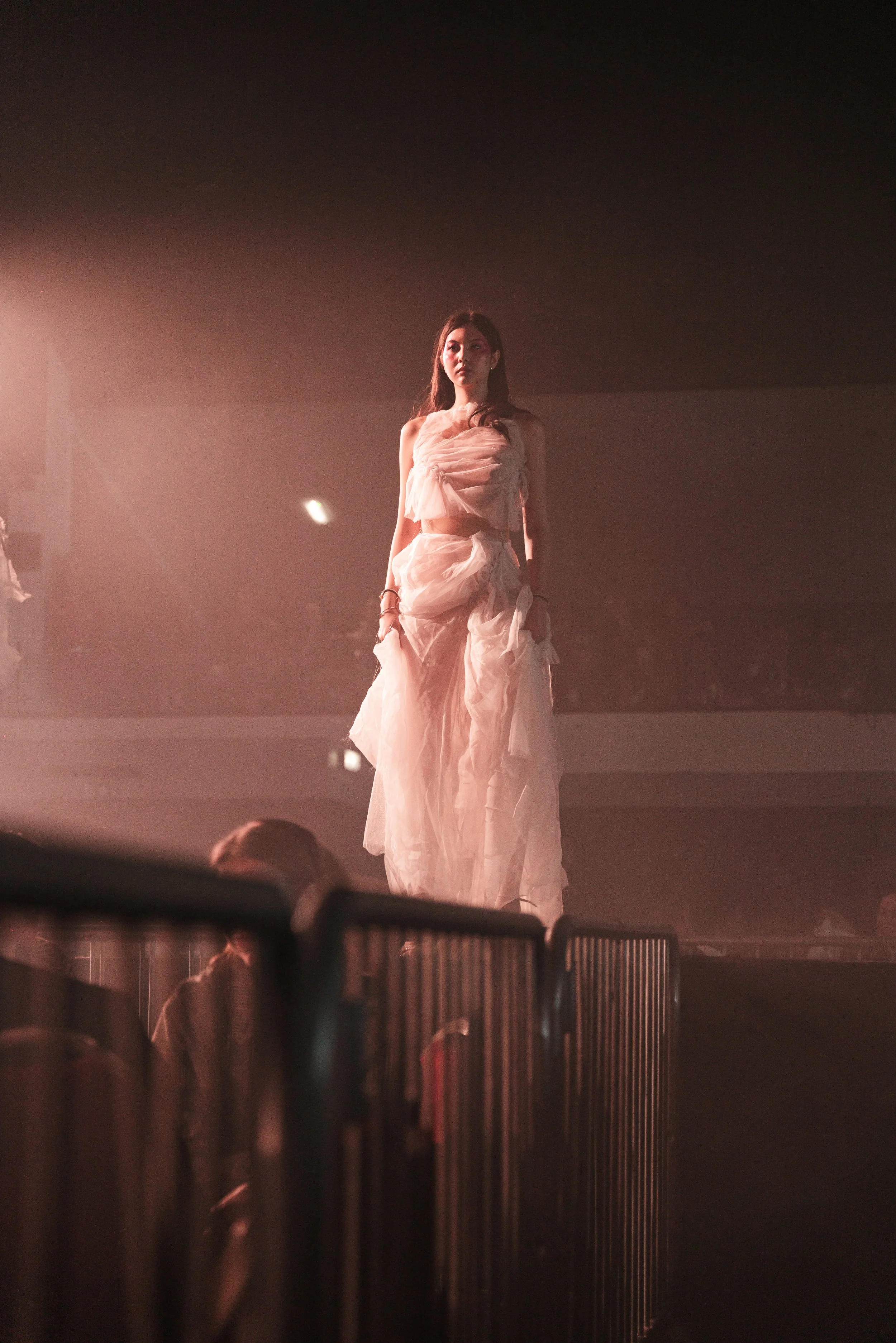 A woman in a flowing, beige dress stands on a stage illuminated by pinkish light, with a dark background and a metal barrier in the foreground.