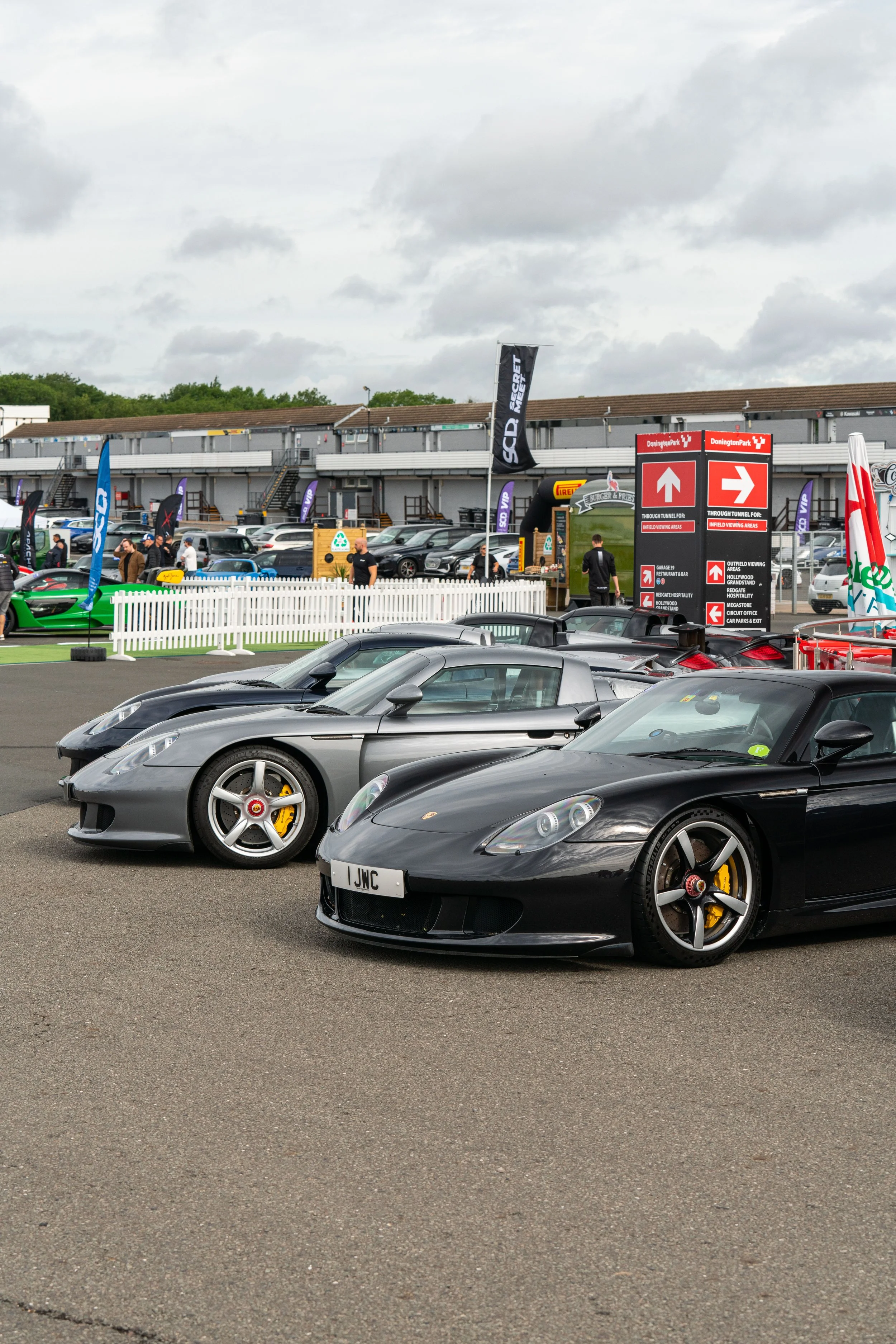 Several luxury sports cars, including a black Porsche and a silver Porsche, parked at an outdoor car event with a race track and parking area in the background.