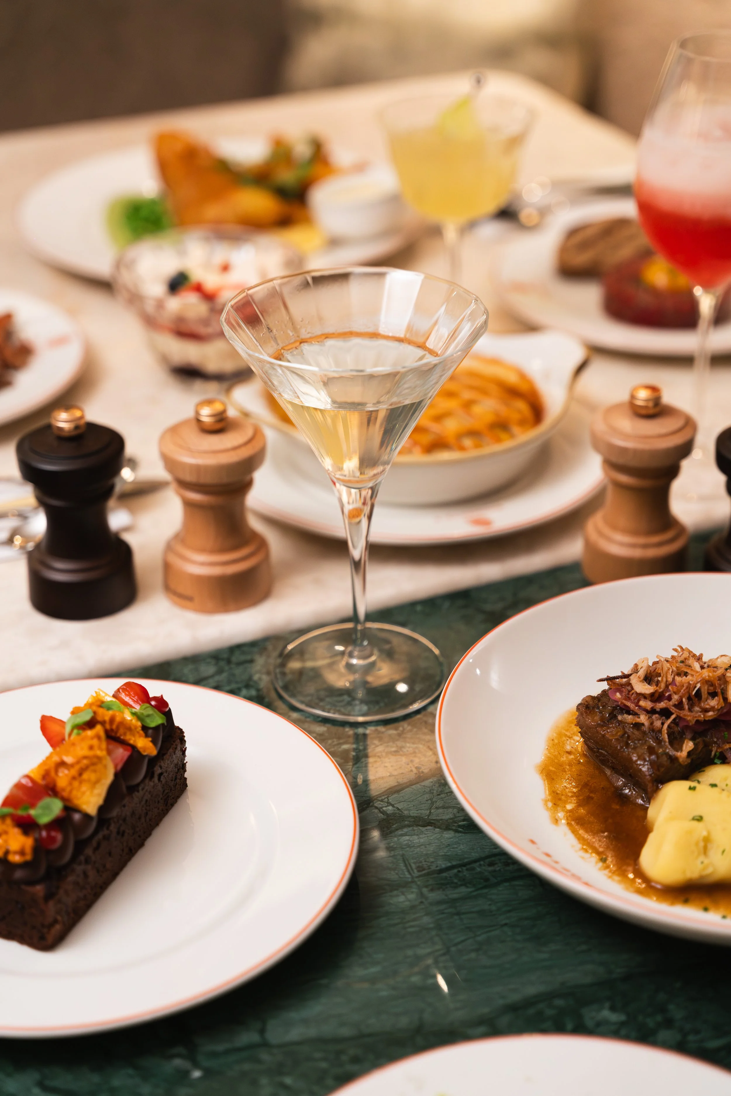 A dining table set with various dishes including a slice of chocolate cake with strawberries and orange toppings, a beef dish with mashed potatoes, a plate of mixed vegetables, a bowl of soup, and a variety of drinks including cocktails, beer, and wa