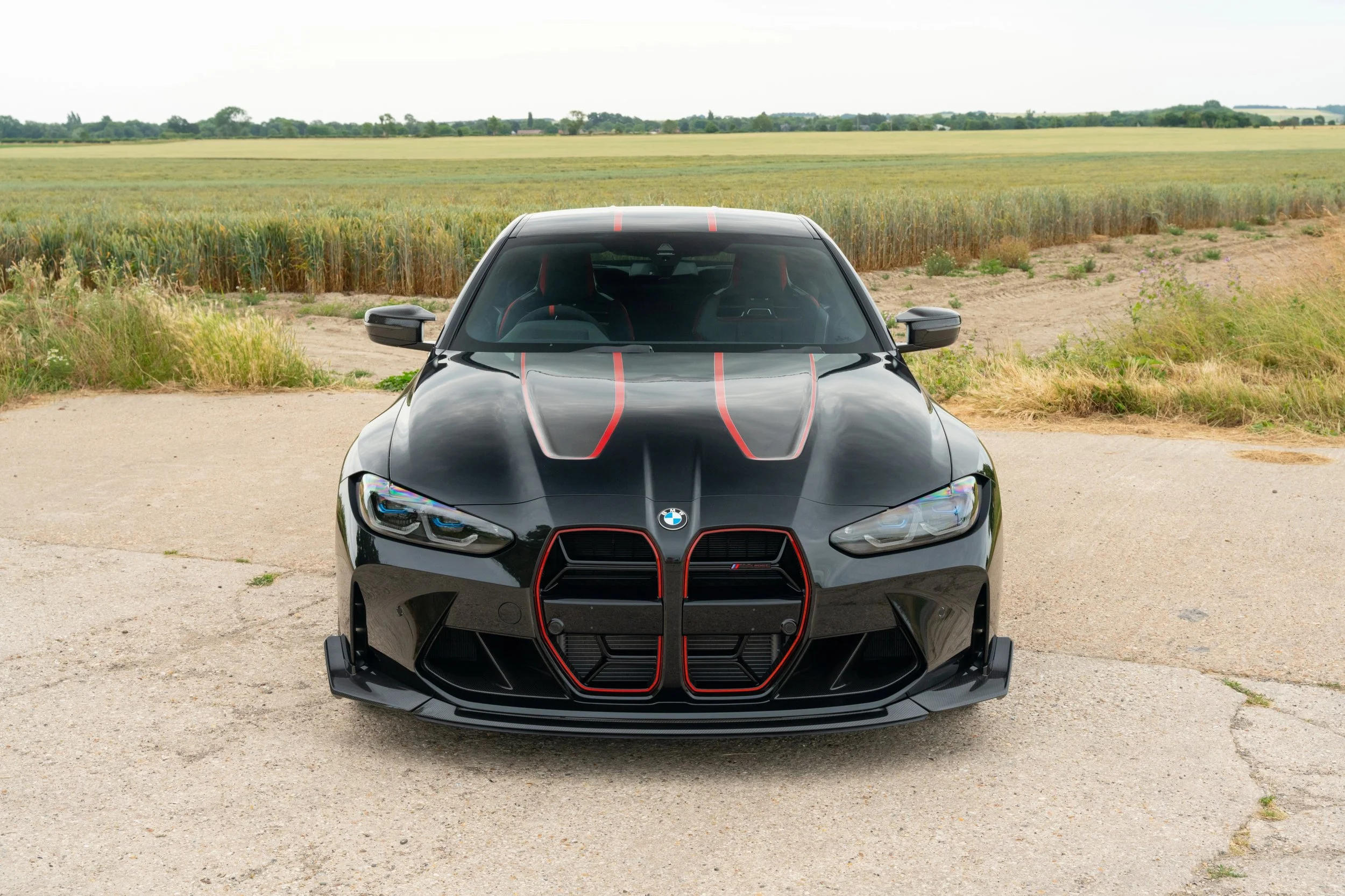 Front view of a black BMW M4 sports car with red accents parked on a concrete surface with a rural field background.