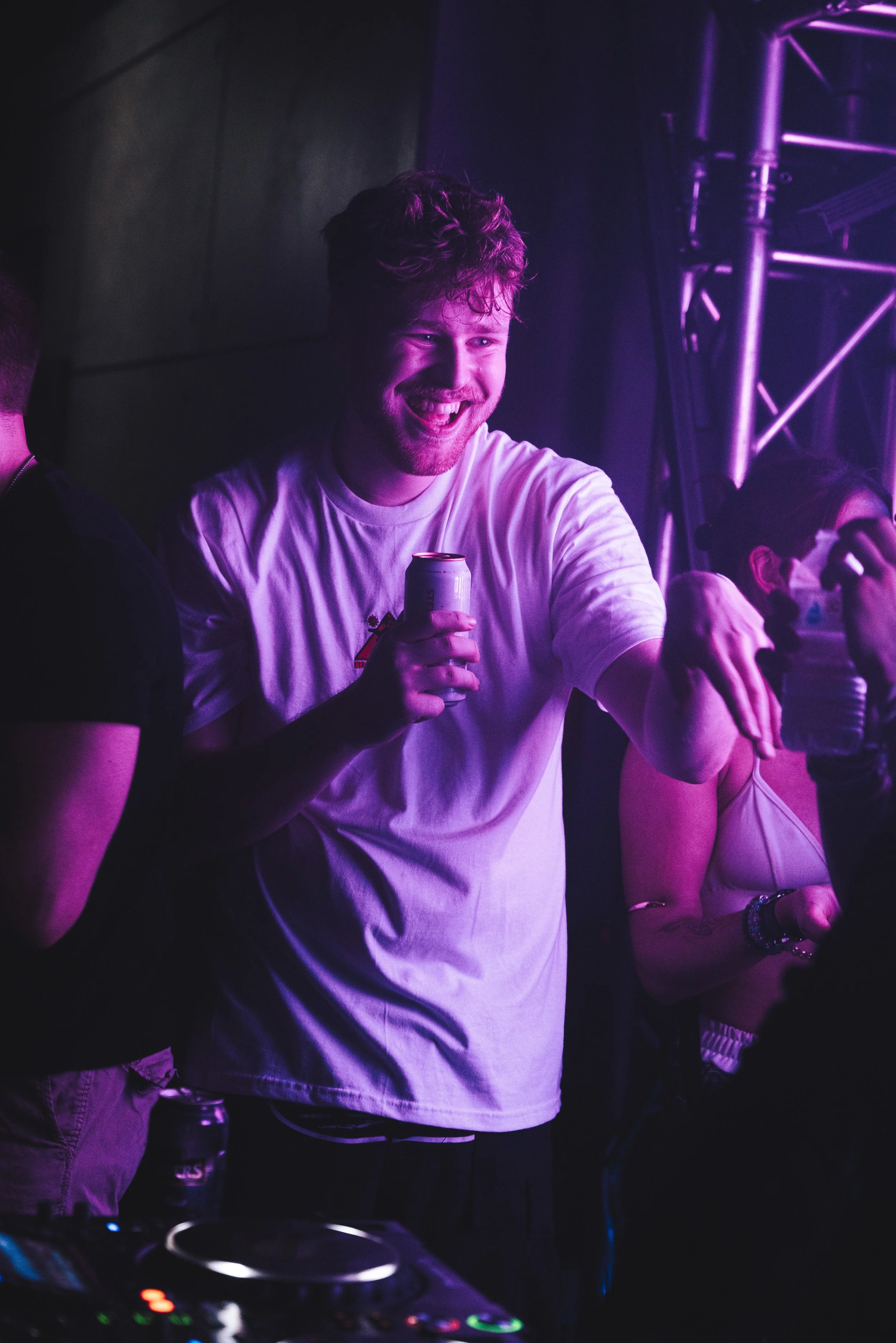 Young man with curly hair smiling and holding a drink at a party or club, illuminated by purple lighting.
