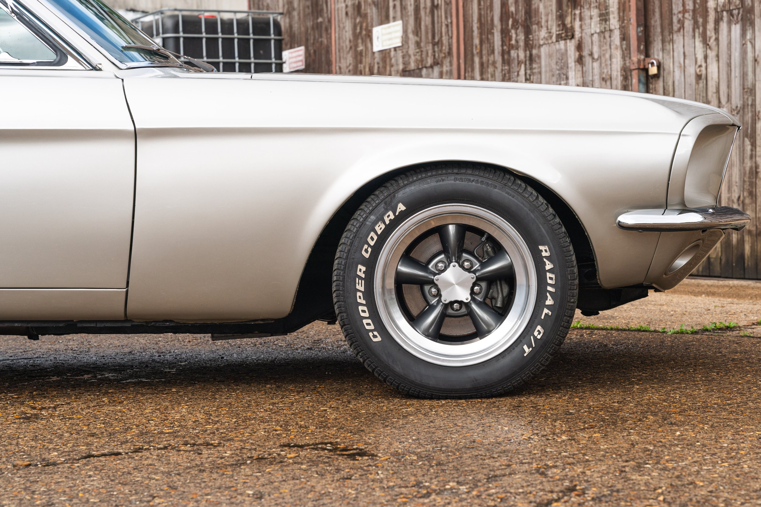 Close-up of a vintage car's front right side, including the wheel with black tire and black rim, and part of the silver body, parked on a wet pavement in front of a wooden fence.