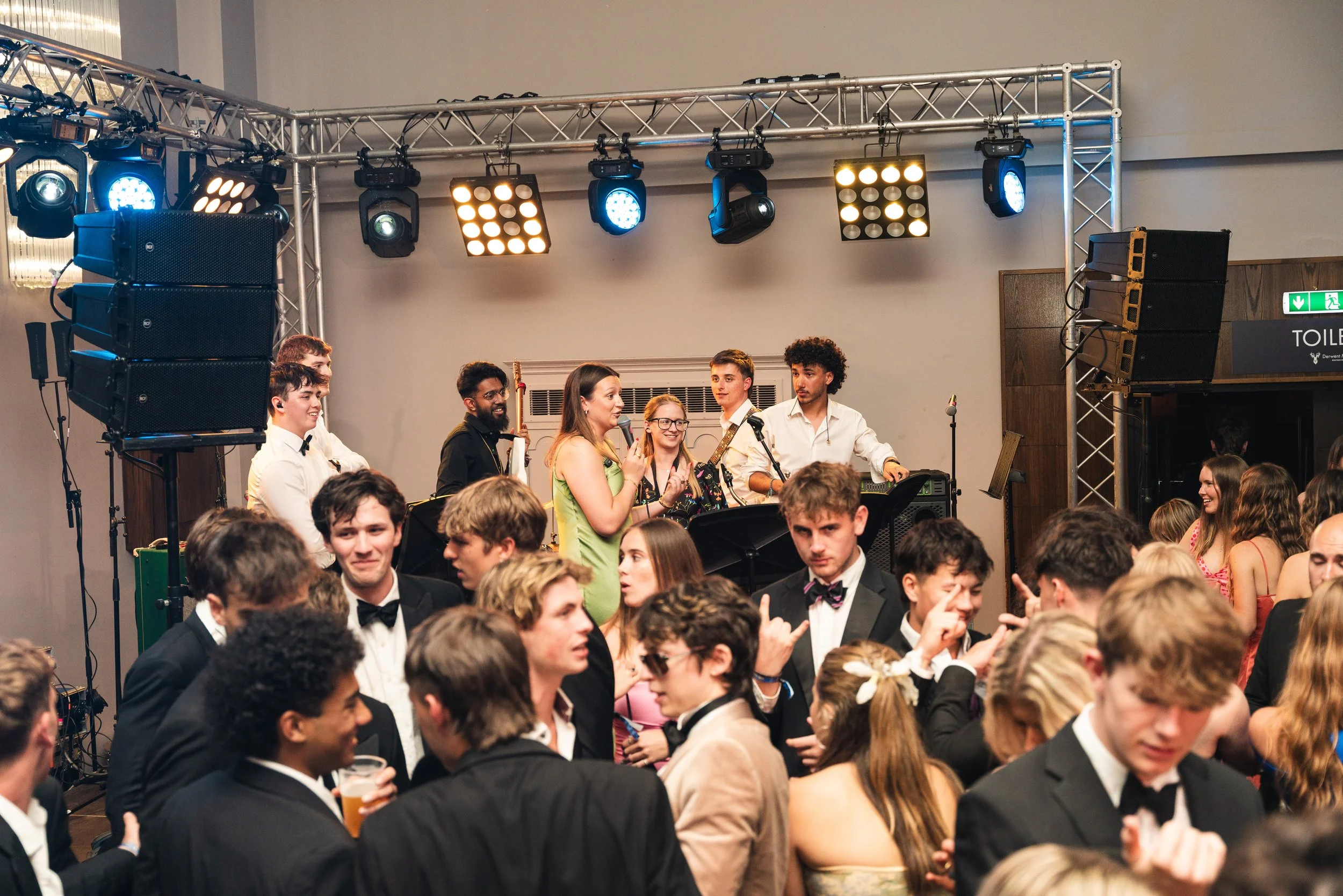 Crowd of people dressed in formal attire dancing and socializing at a party or event with a live band playing on stage in the background.