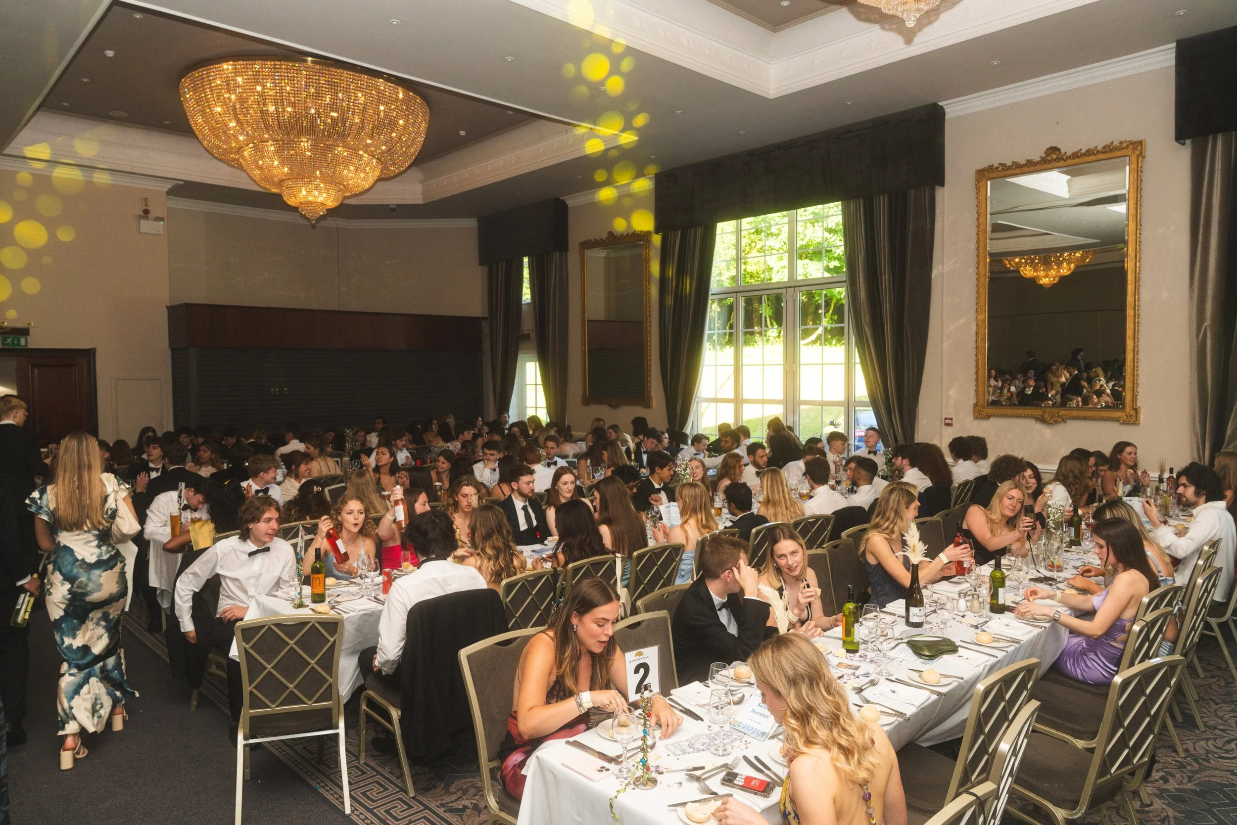 A large banquet hall filled with formally dressed people seated at long tables, celebrating an event with plates of food and drinks, illuminated by a grand chandelier, with large windows letting in natural light, and framed mirrors on the walls.