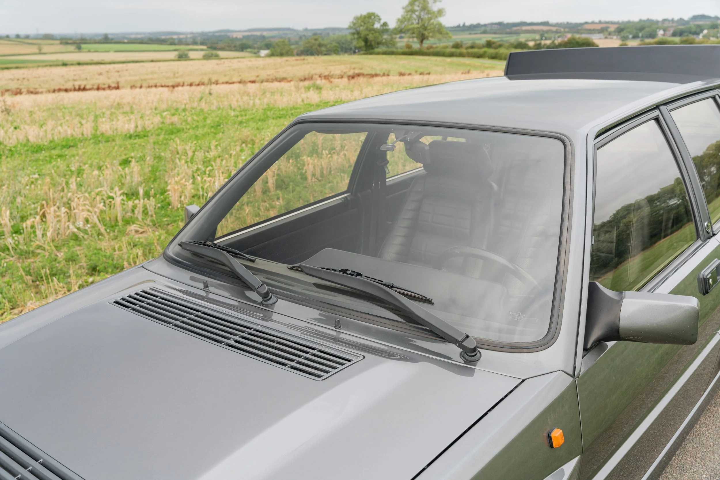 Silver vintage hatchback car parked in a rural field with green and yellow fields and trees in the background.