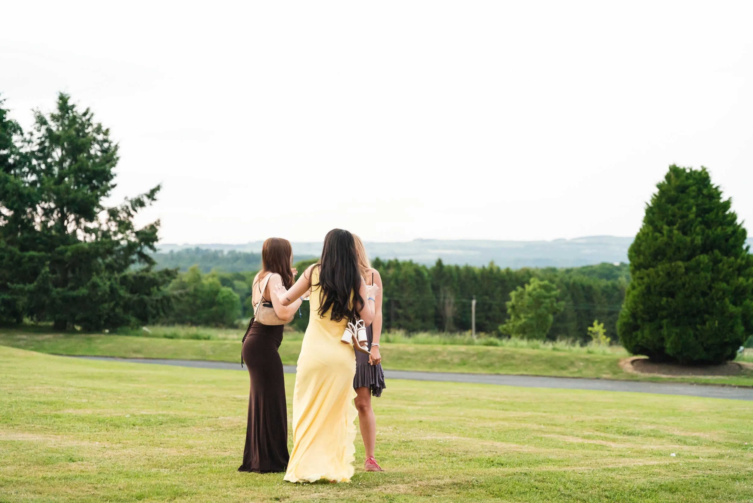 Three women standing together outdoors on a grassy field, with trees and hills in the background.