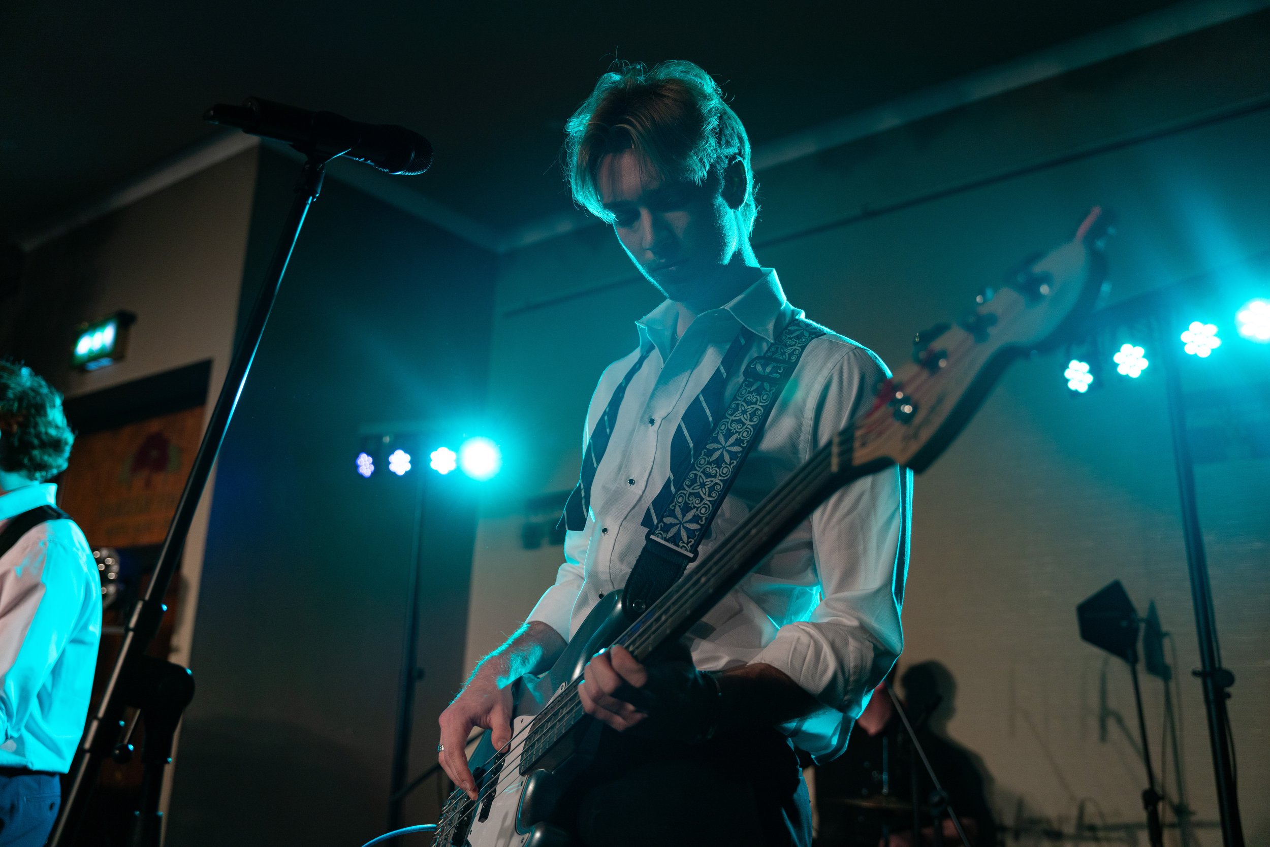 A young male musician playing an electric bass guitar on stage under blue and purple stage lights, wearing a white shirt and suspenders.