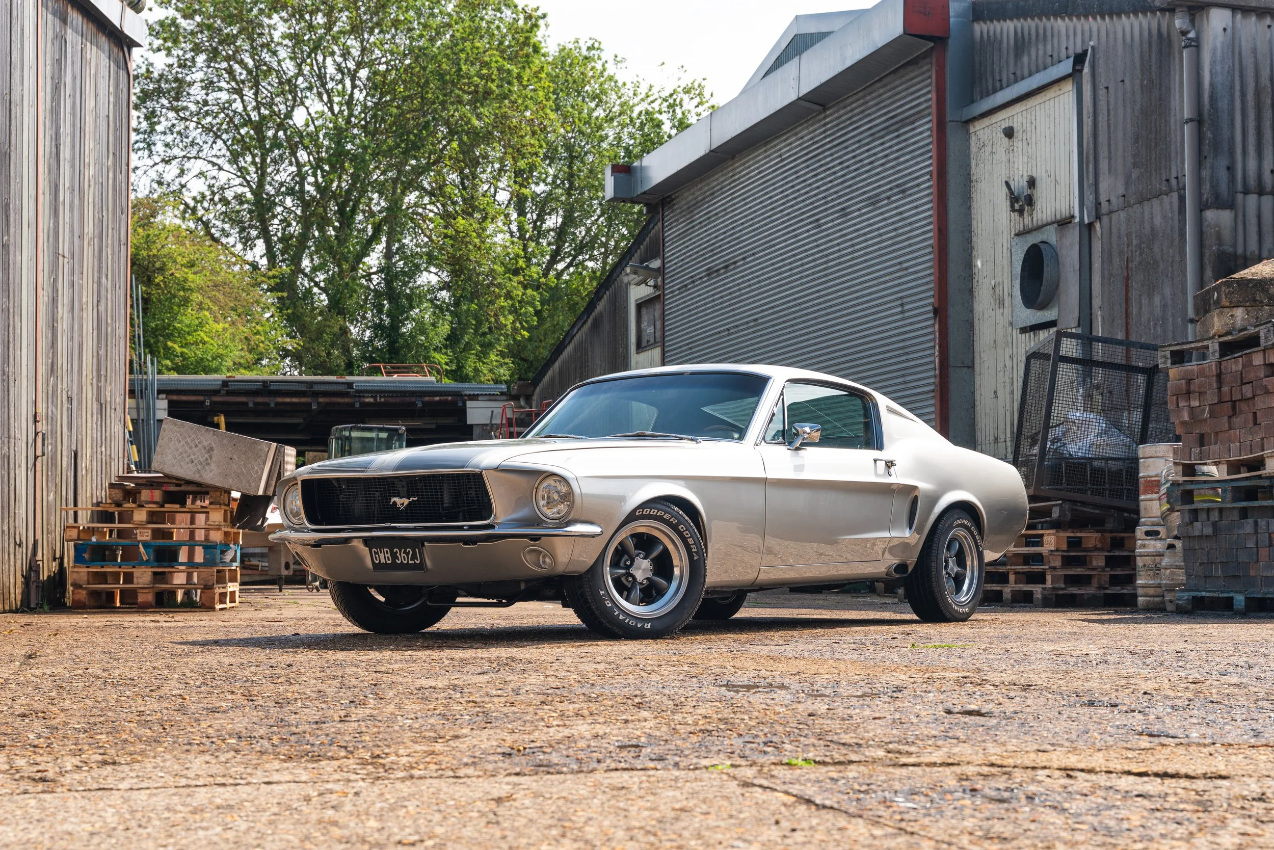 Silver classic Ford Mustang car parked outside near industrial buildings and wood pallets.
