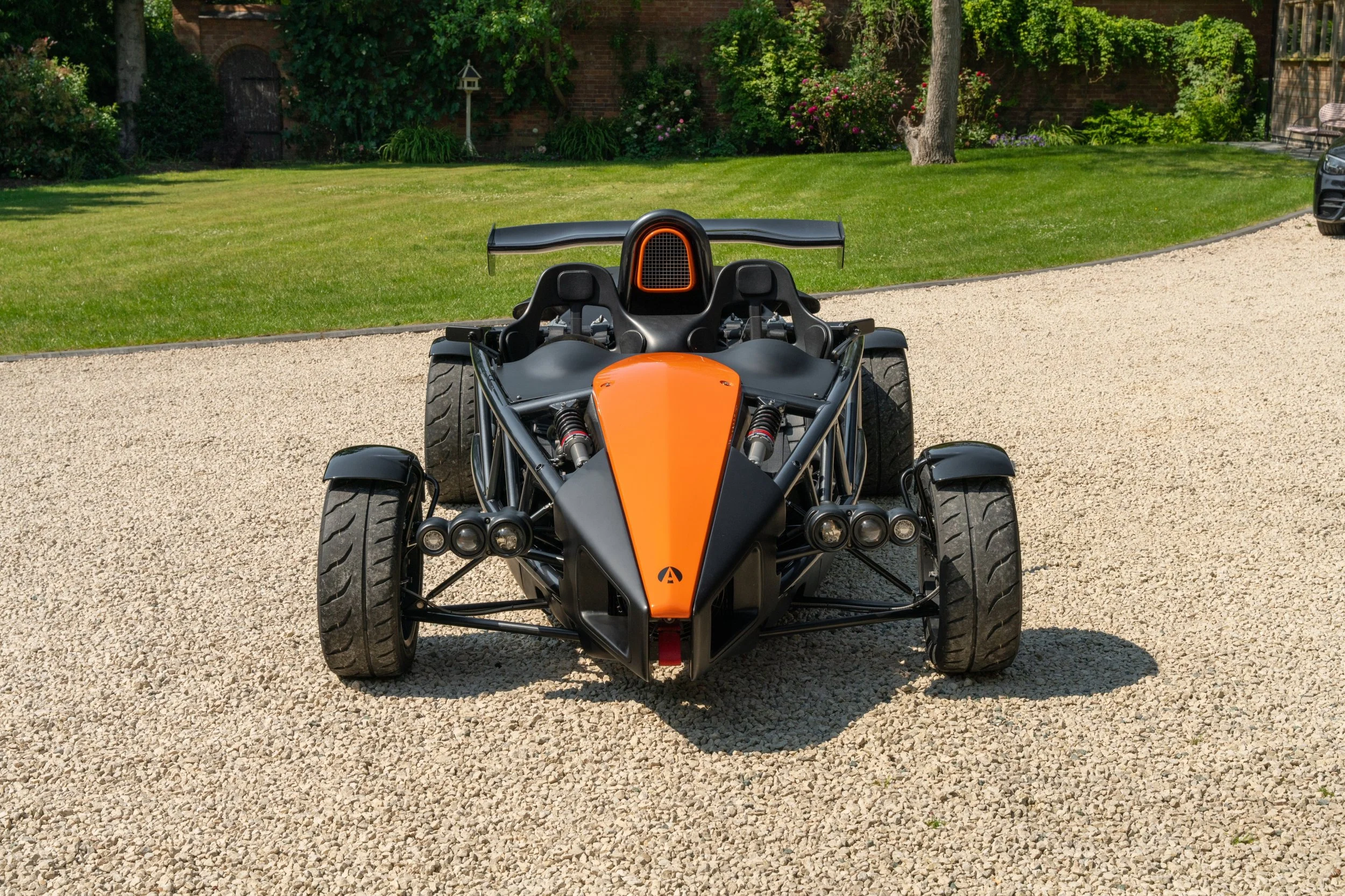 Front view of a black and orange three-wheeled sports car on a gravel driveway with a grassy yard and trees in the background.