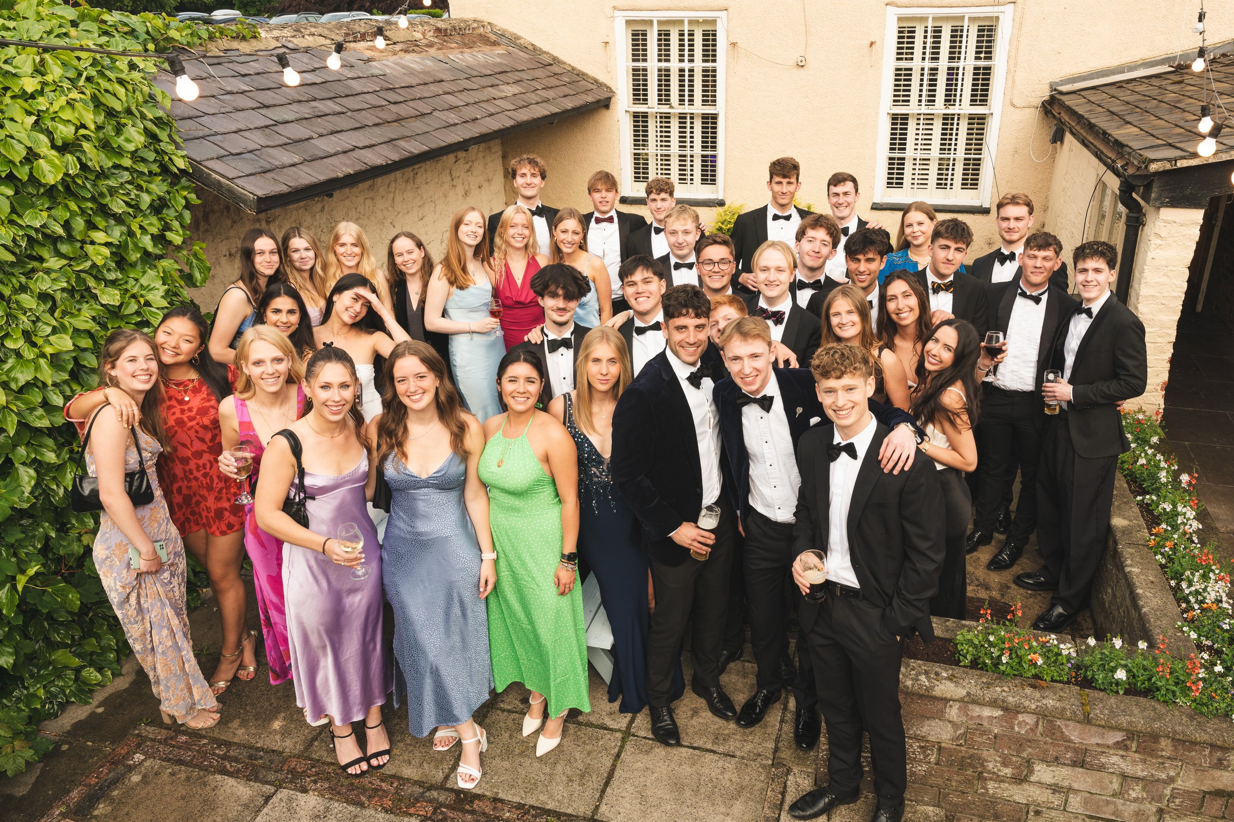 A large group of young adults dressed in formal attire, celebrating at an outdoor gathering in the evening with string lights overhead. The group is smiling and posing for a photo in a courtyard with lush greenery and a building with white windows in