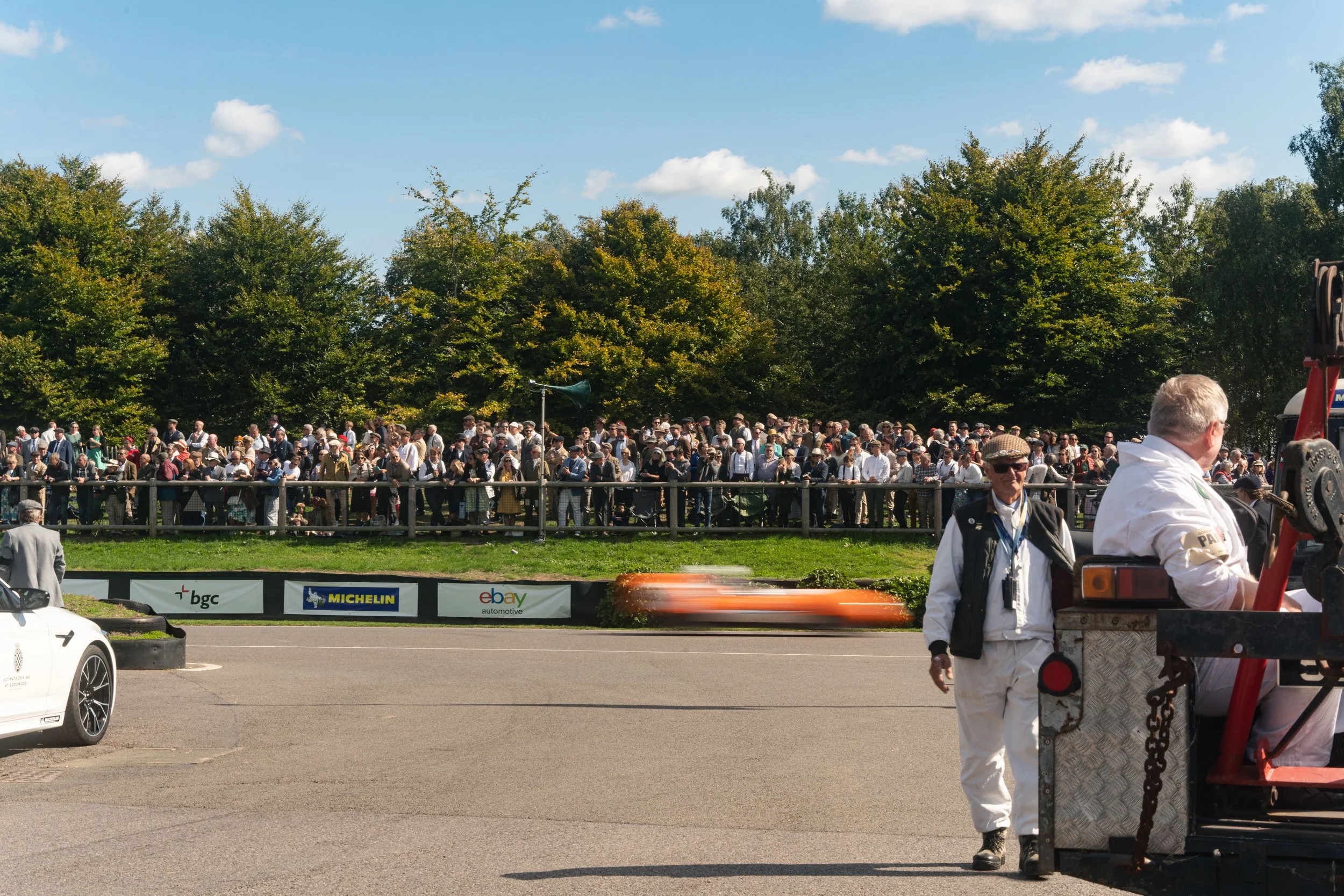 A large crowd of people gathered behind a safety barrier at a racing event, with two men in white uniforms and event staff equipment in the foreground. The scene is outdoors on a sunny day with a clear blue sky and green trees in the background.