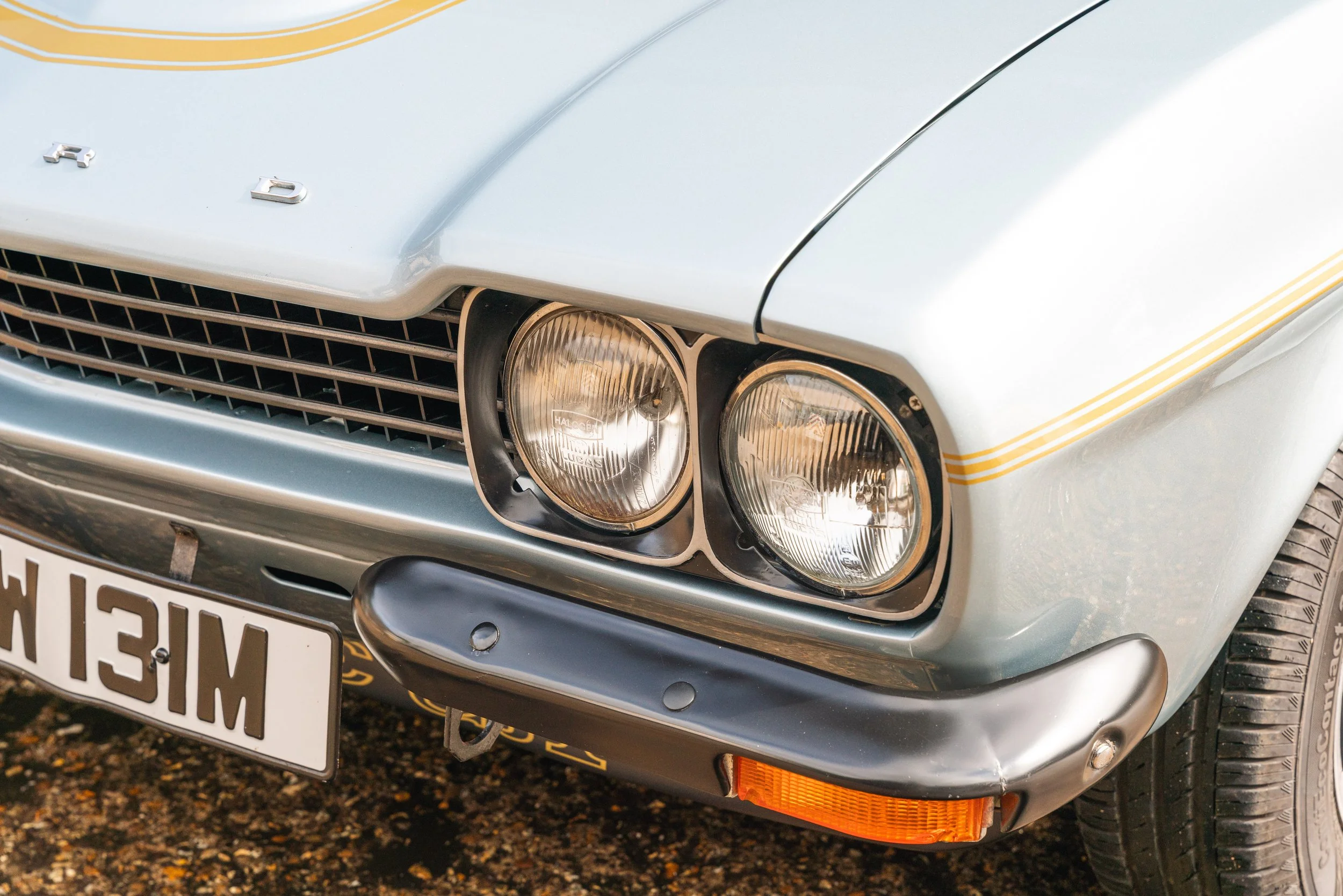 Close-up of a vintage light blue Ford car's front end, showing dual round headlights, grille, front bumper with an orange turn signal, and part of the front wheel.