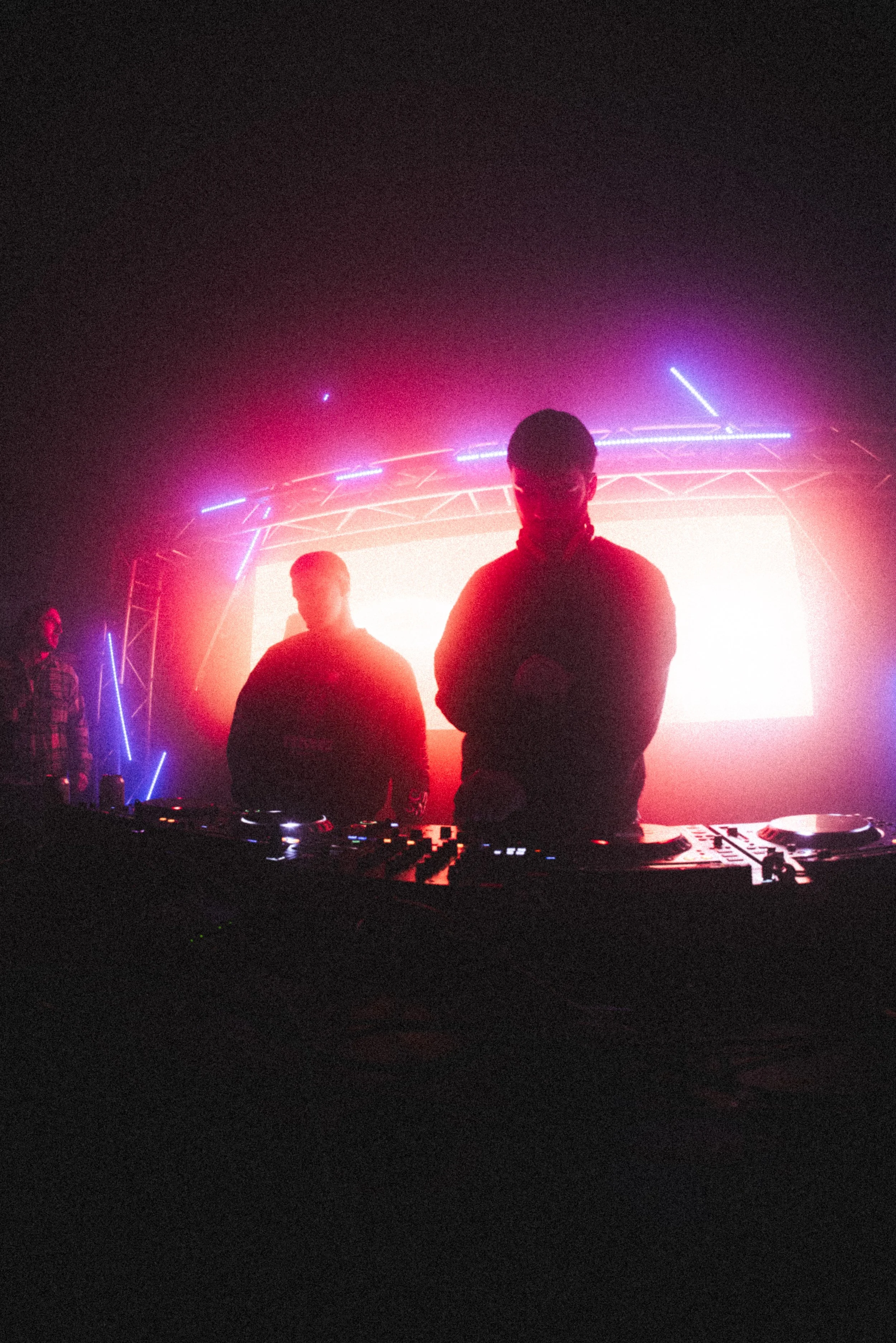 Dark scene of a DJ performing on stage with neon lights and large bright screen behind him, with silhouettes of other people around.
