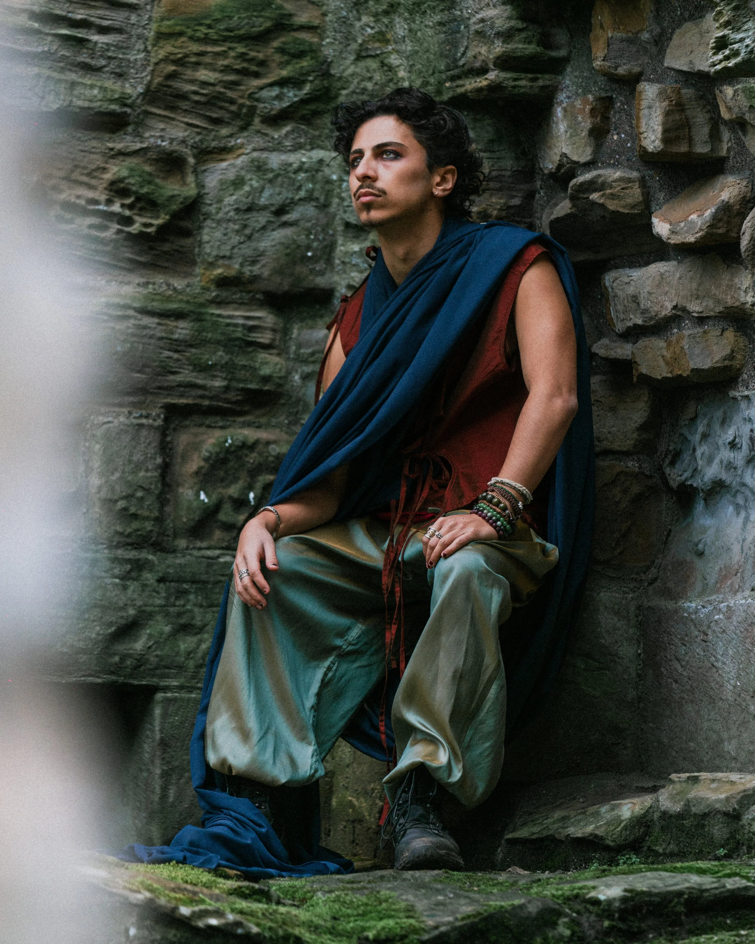A person in historical clothing with a blue cloak, brown sleeveless top, and greenish baggy pants, sitting against a stone wall with moss and small green plants growing on it.