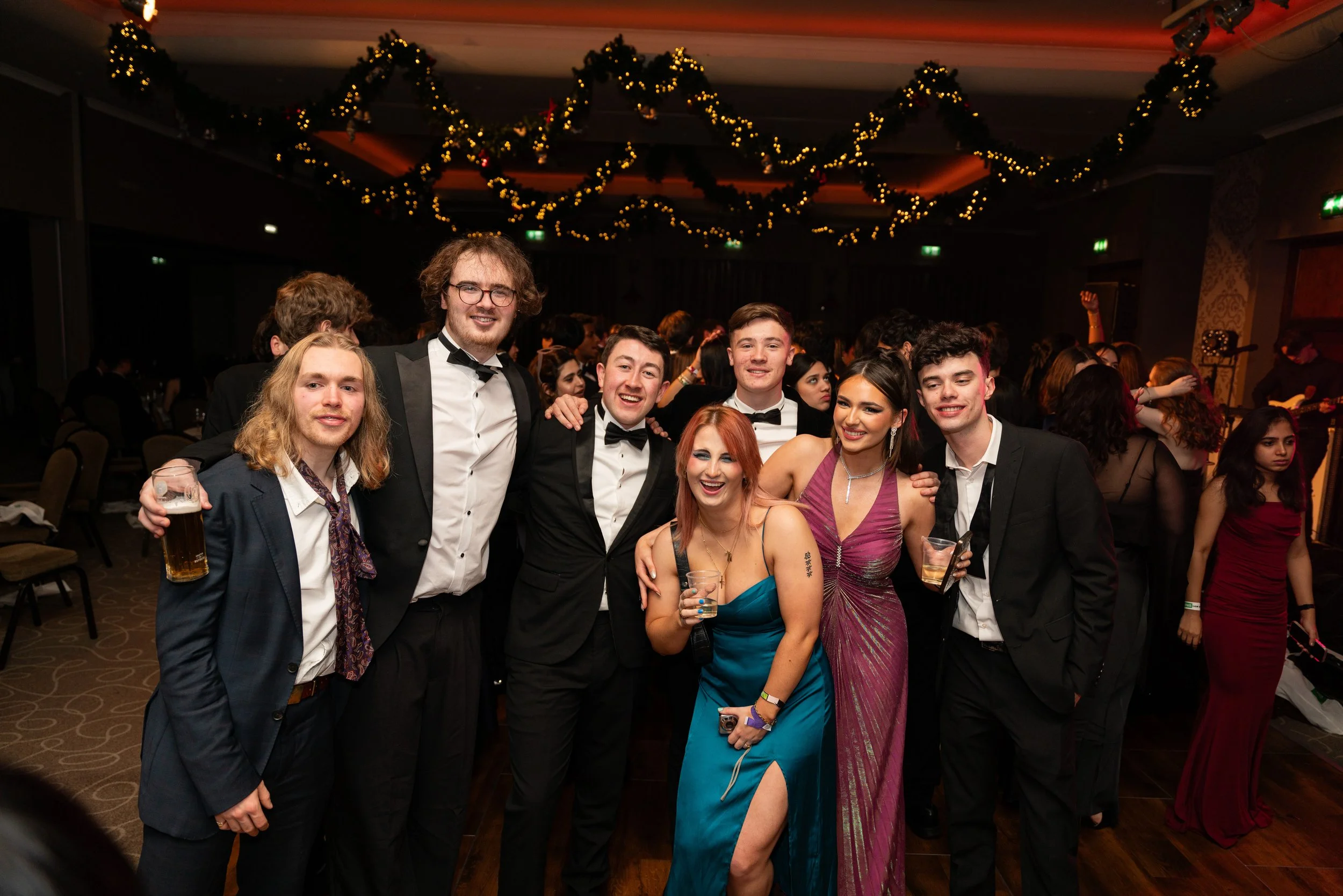 Group of multiple young adults in formal attire at a festive party, smiling and holding drinks, with decorative holiday garlands hanging from the ceiling in the background.