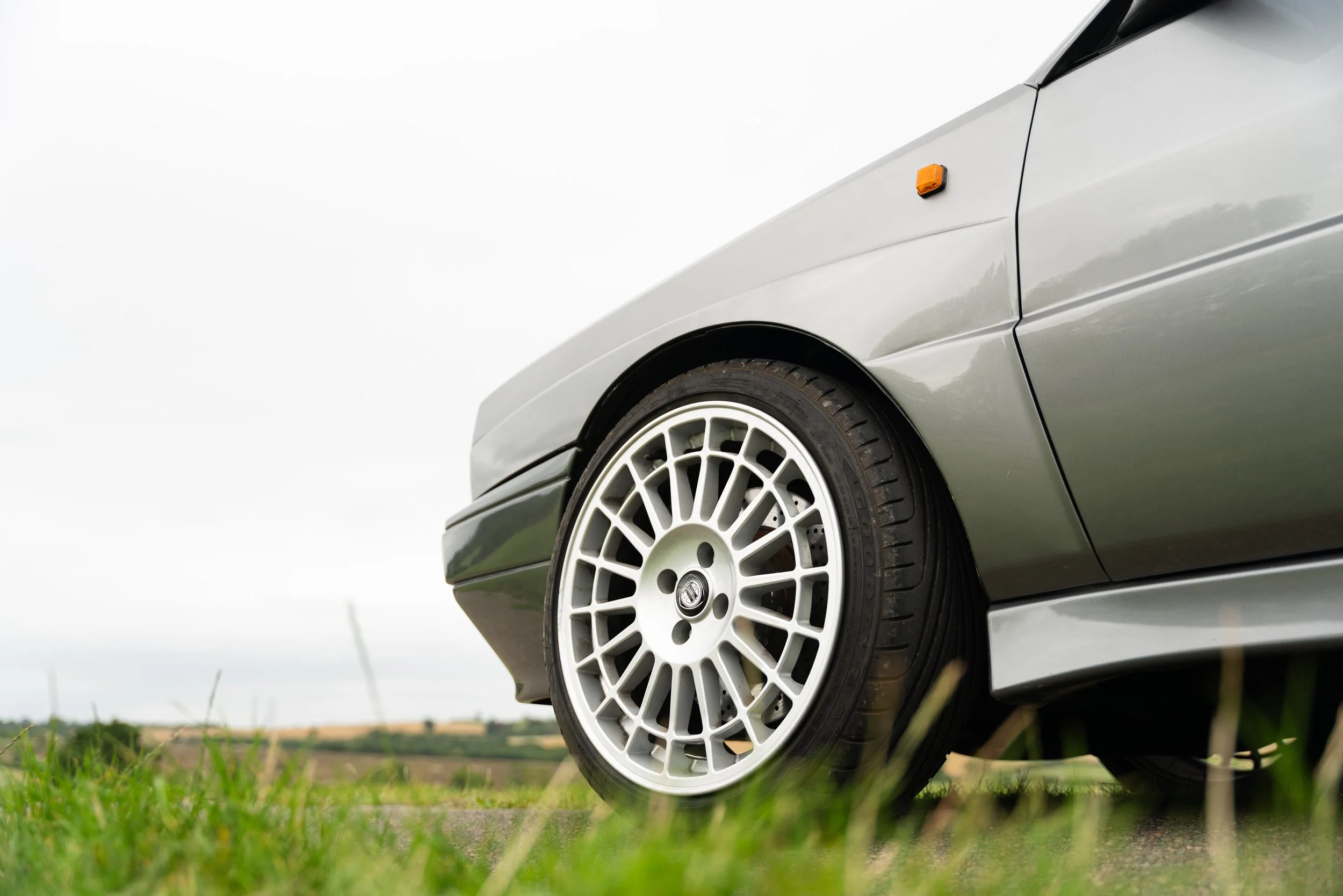 Close-up of a vintage silver car's front right wheel and body, parked on grass with a cloudy sky.