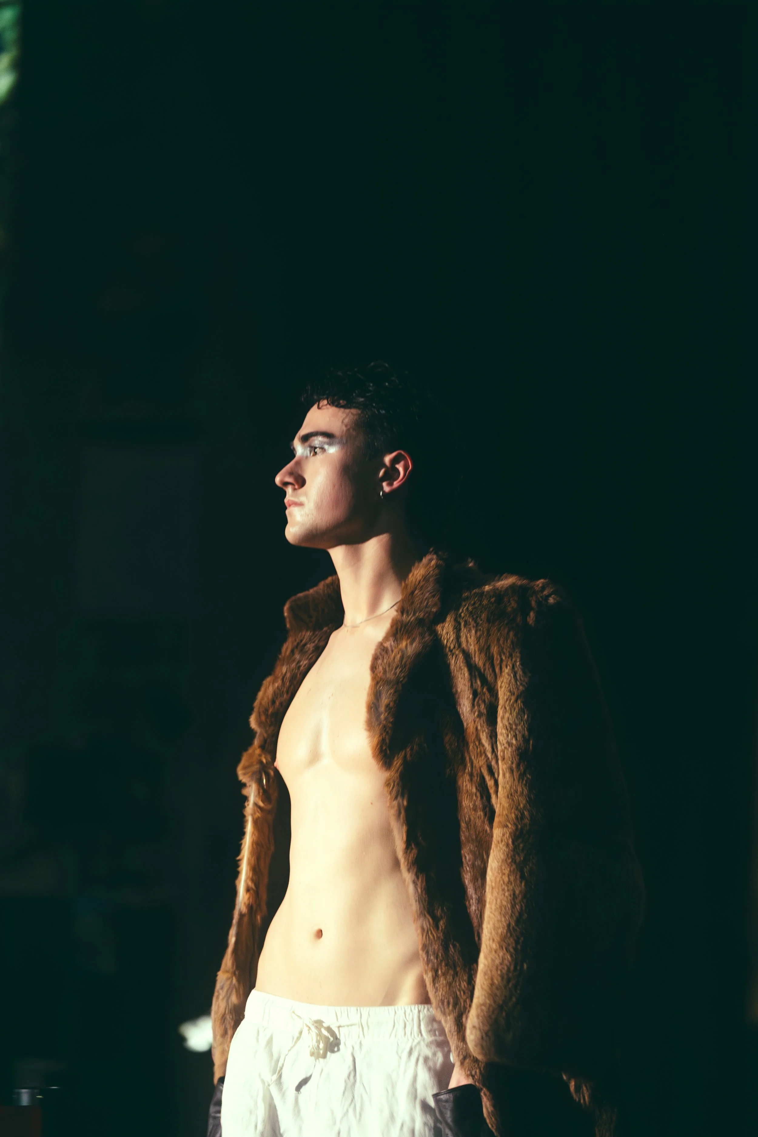 A young man with dark hair and earrings wearing a fur coat, shirtless, and white pants standing in profile against a dark background.