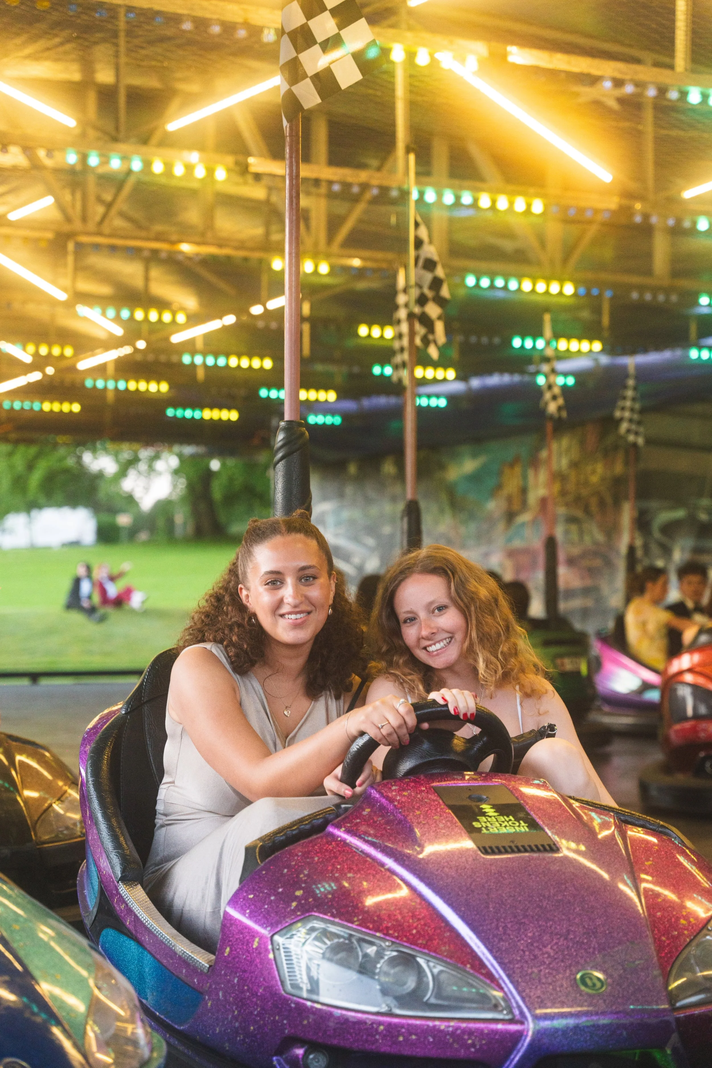 Two young women with curly hair smiling while riding a colorful amusement park bumper car.