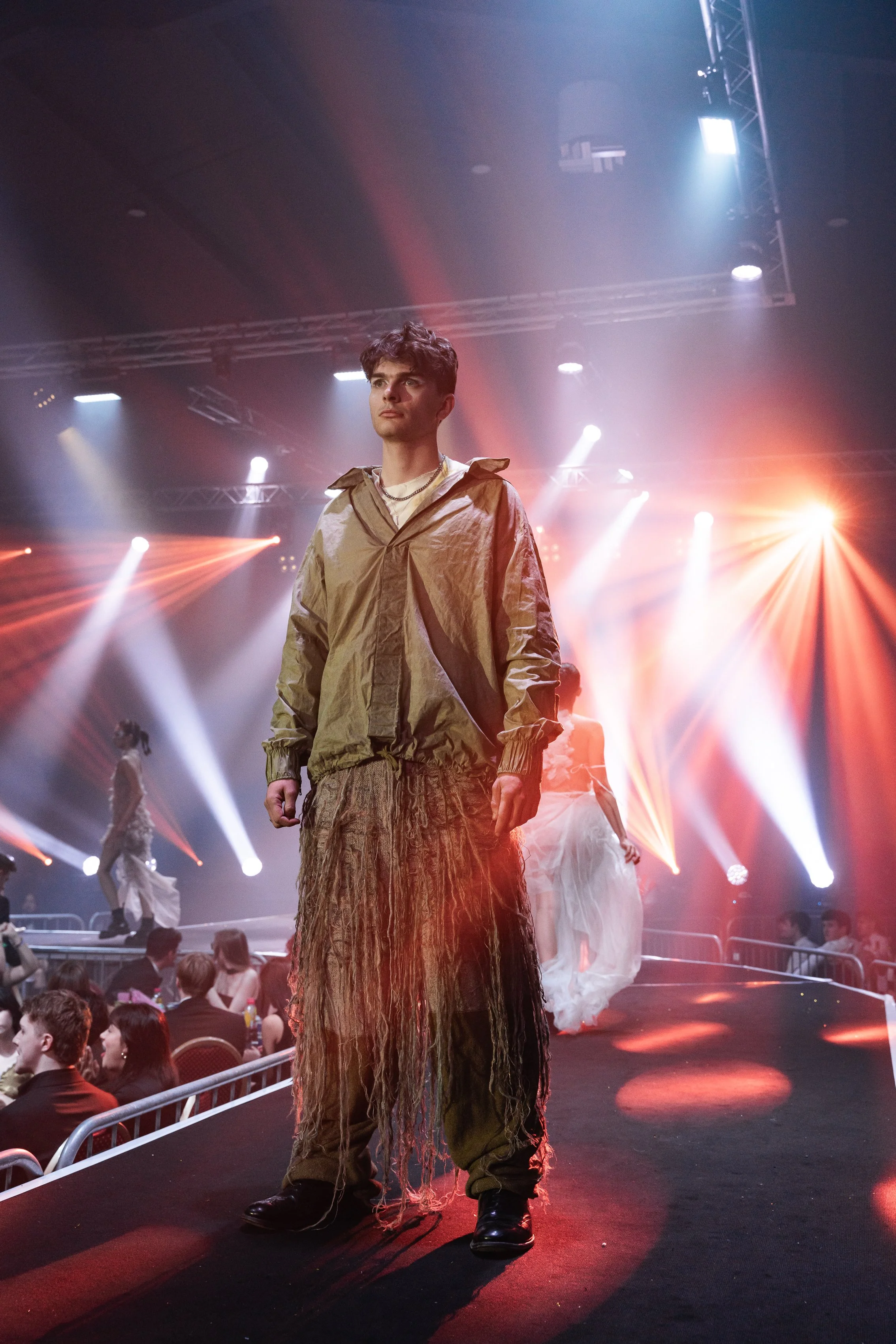 A male model walking down a runway during a fashion show with bright stage lights and an audience in the background.