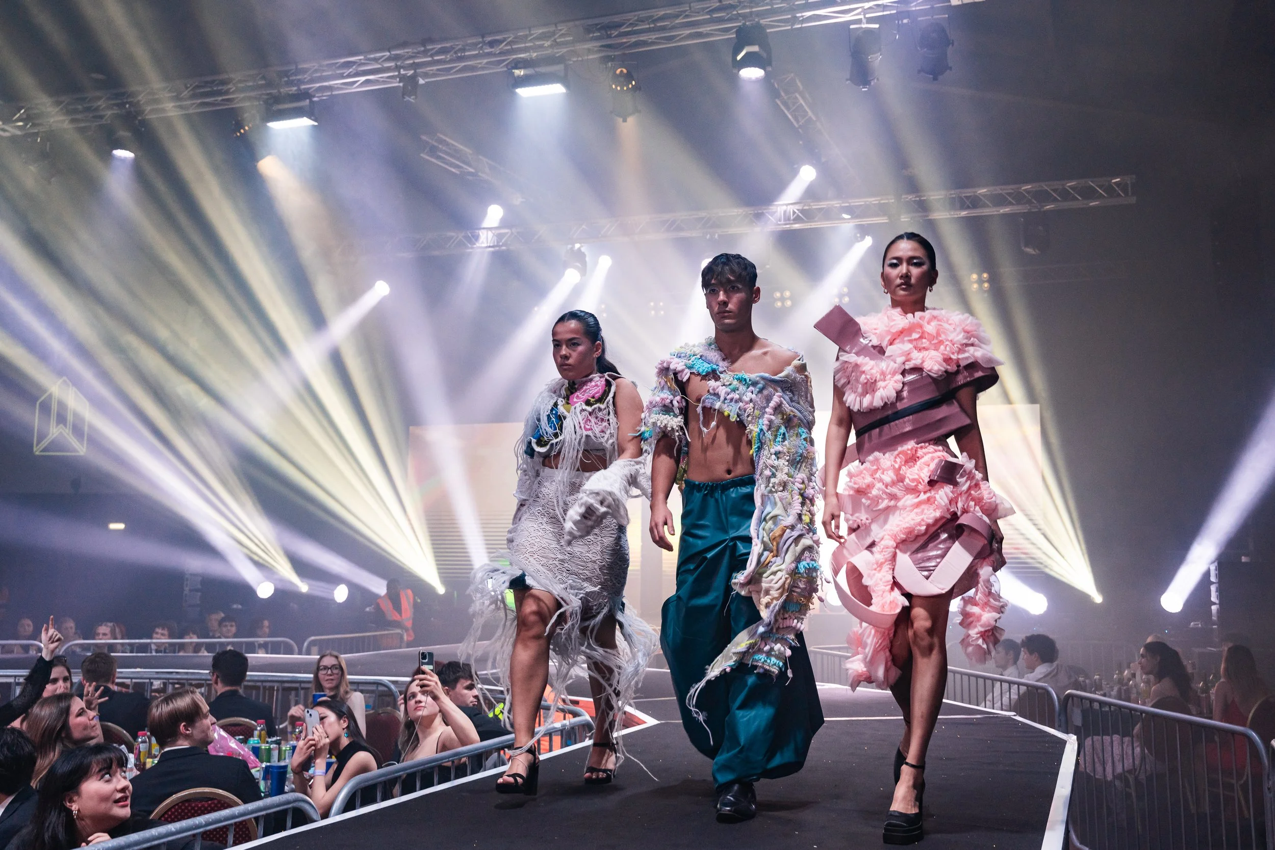Three models walking down a runway at a fashion show, with bright stage lights and audience watching.