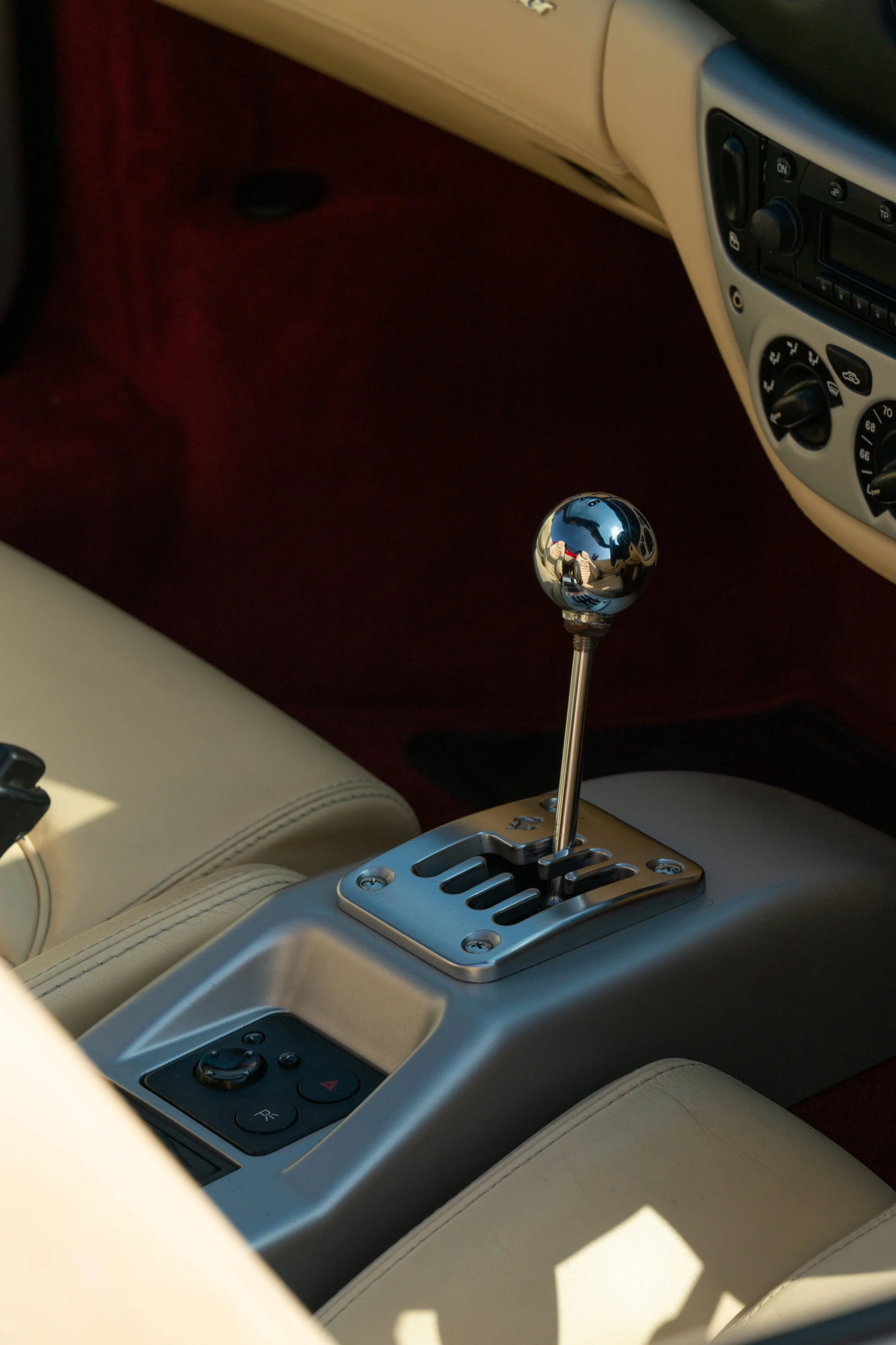 The interior of a luxury car showing a gear shift lever with a shiny metallic ball knob, climate control and radio panel, beige seats, and a red carpeted floor.