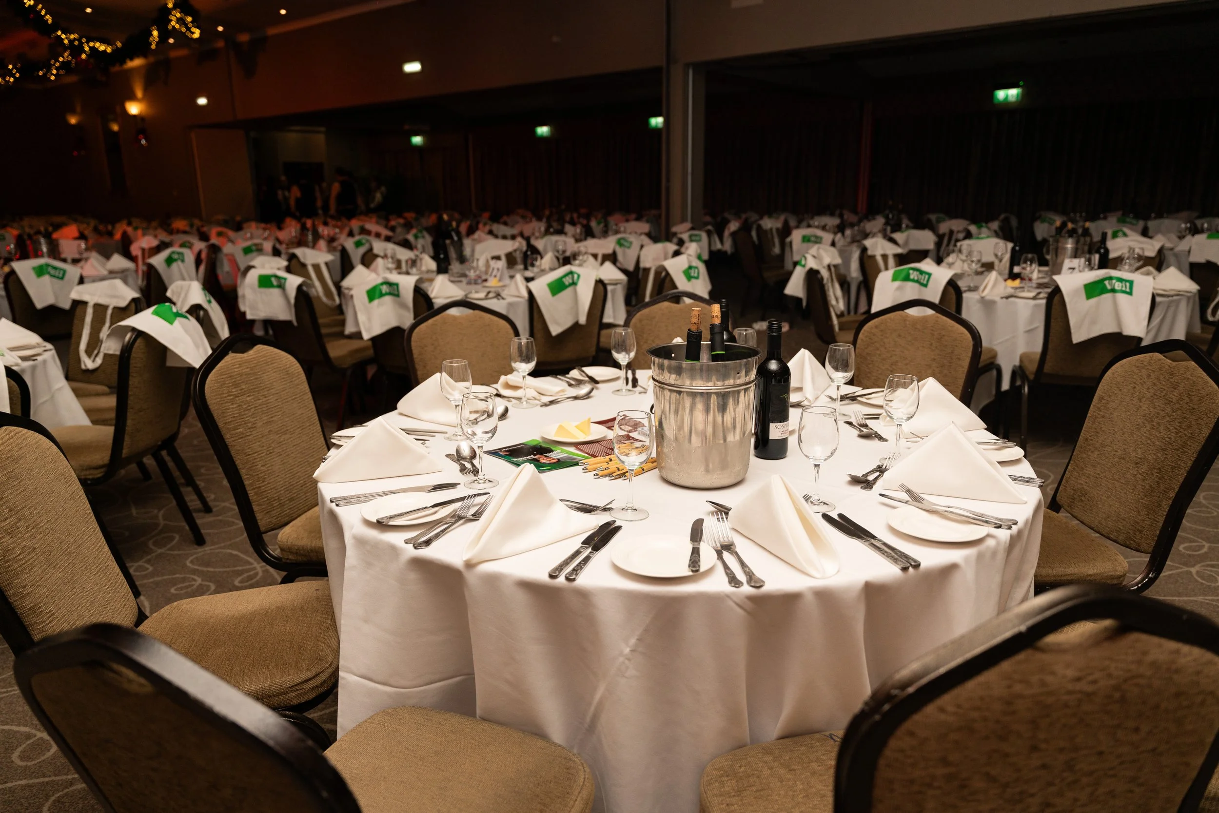 A round banquet table with white tablecloths, set for a formal dinner with white napkins, glassware, silverware, and small plates, in a large event hall decorated for a celebration.