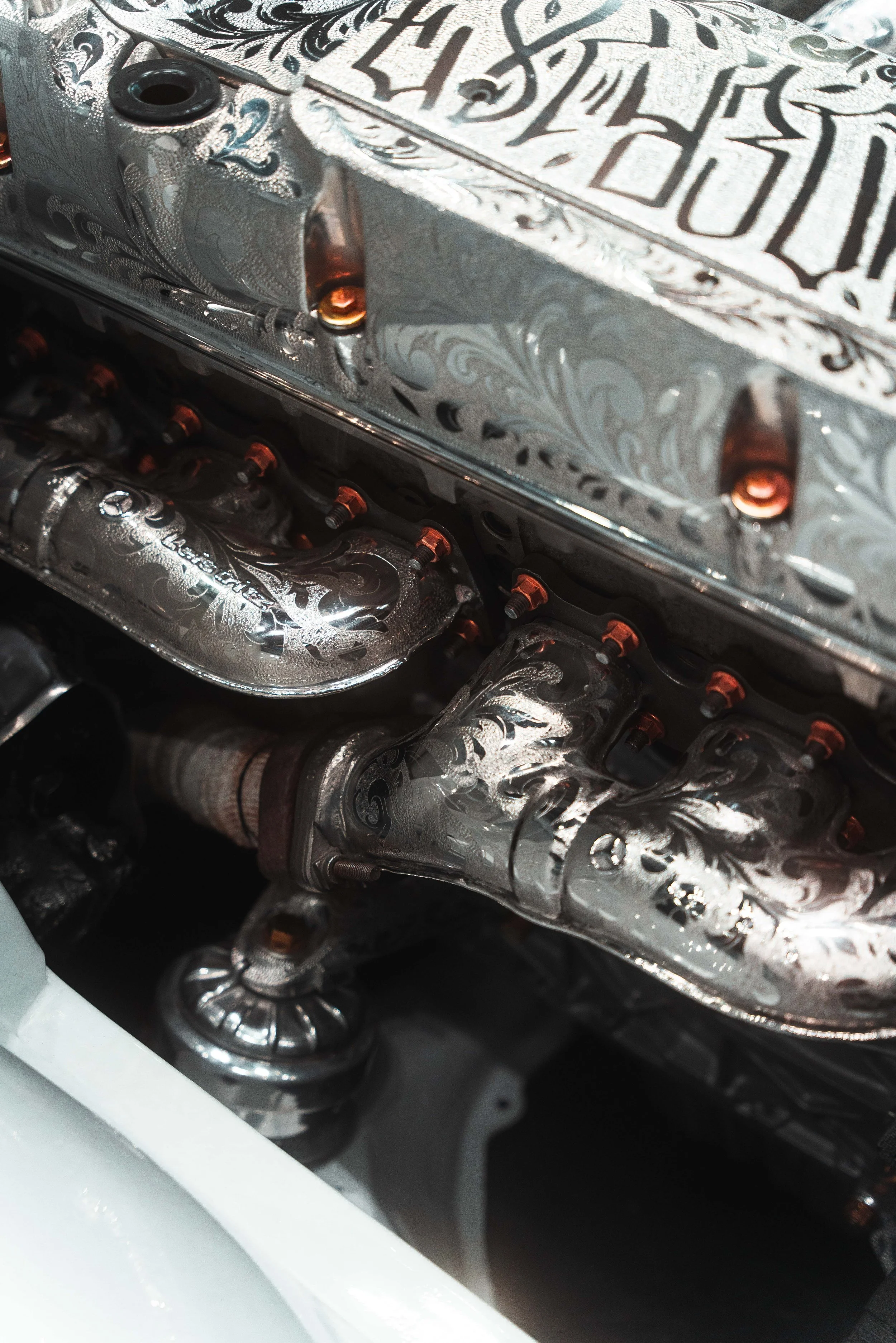 Close-up of a highly detailed, engraved metal engine part with intricate floral and swirling patterns, featuring multiple bolts and a polished finish.