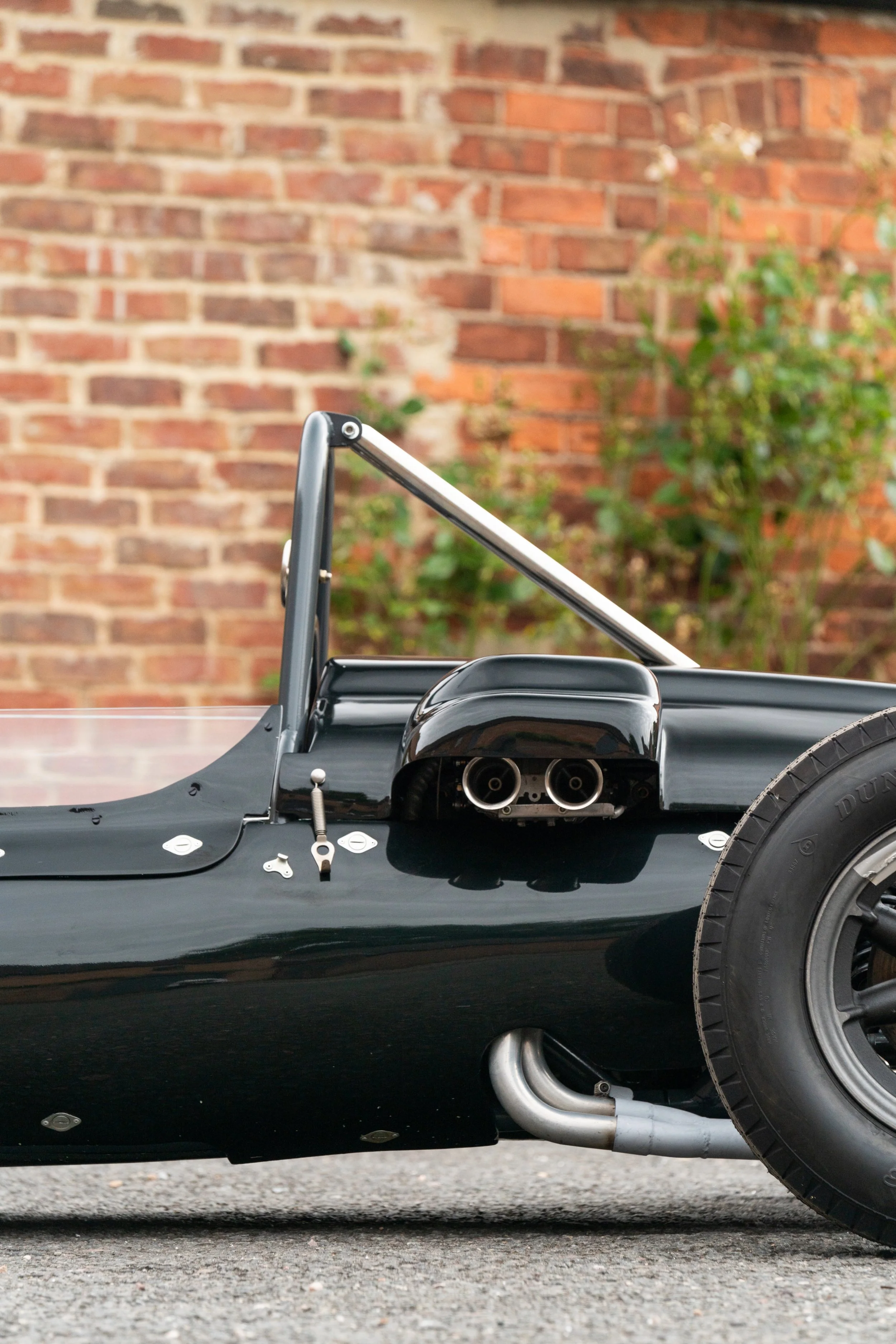 Close-up of the side of a vintage racing car with black bodywork, showing exhaust pipes, a suspension component, and a rear tire, with a brick wall and greenery in the background.