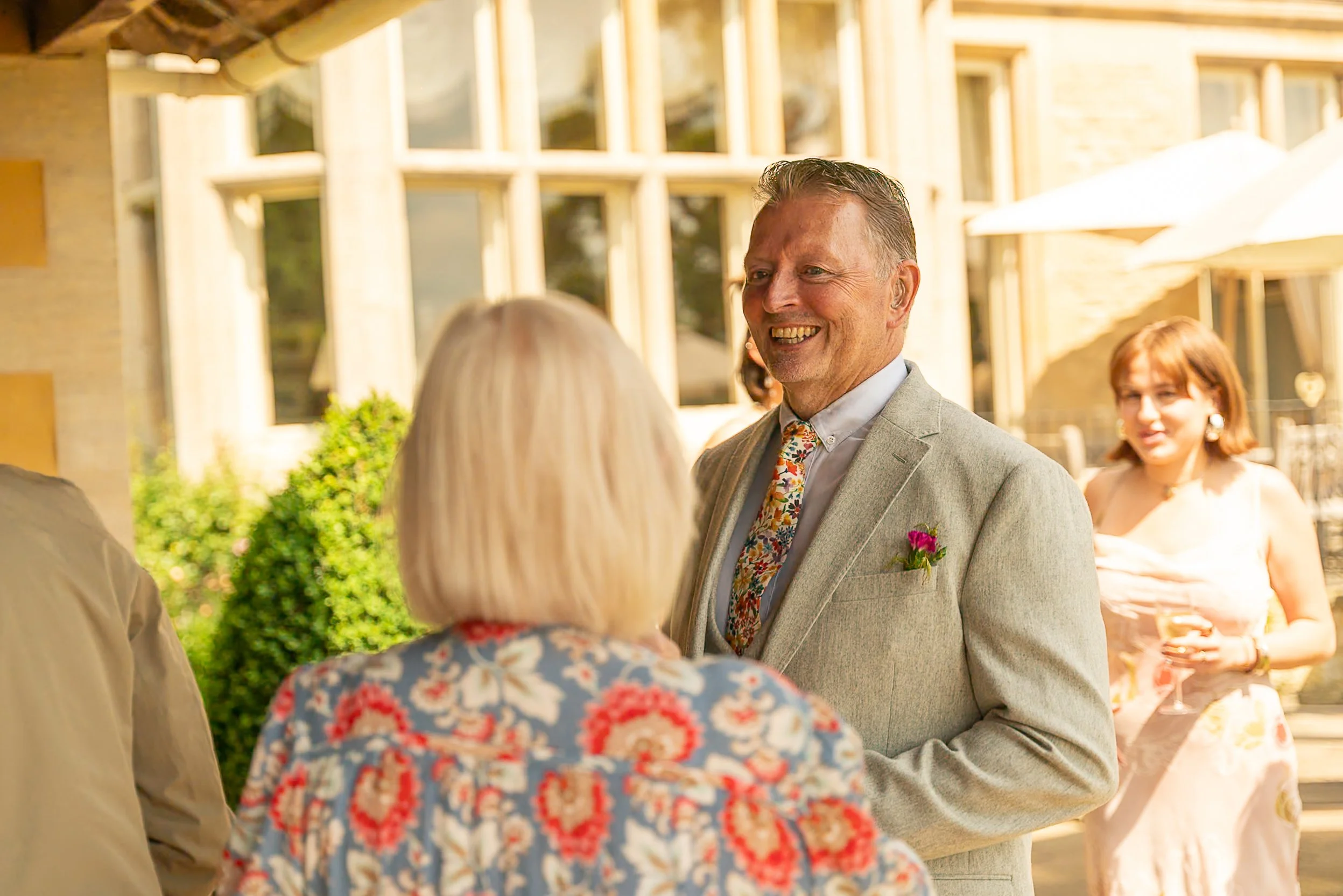 A man smiling and talking to an older woman at an outdoor event, with a woman in a beige dress holding a glass in the background, sunny weather, and a building with large windows.