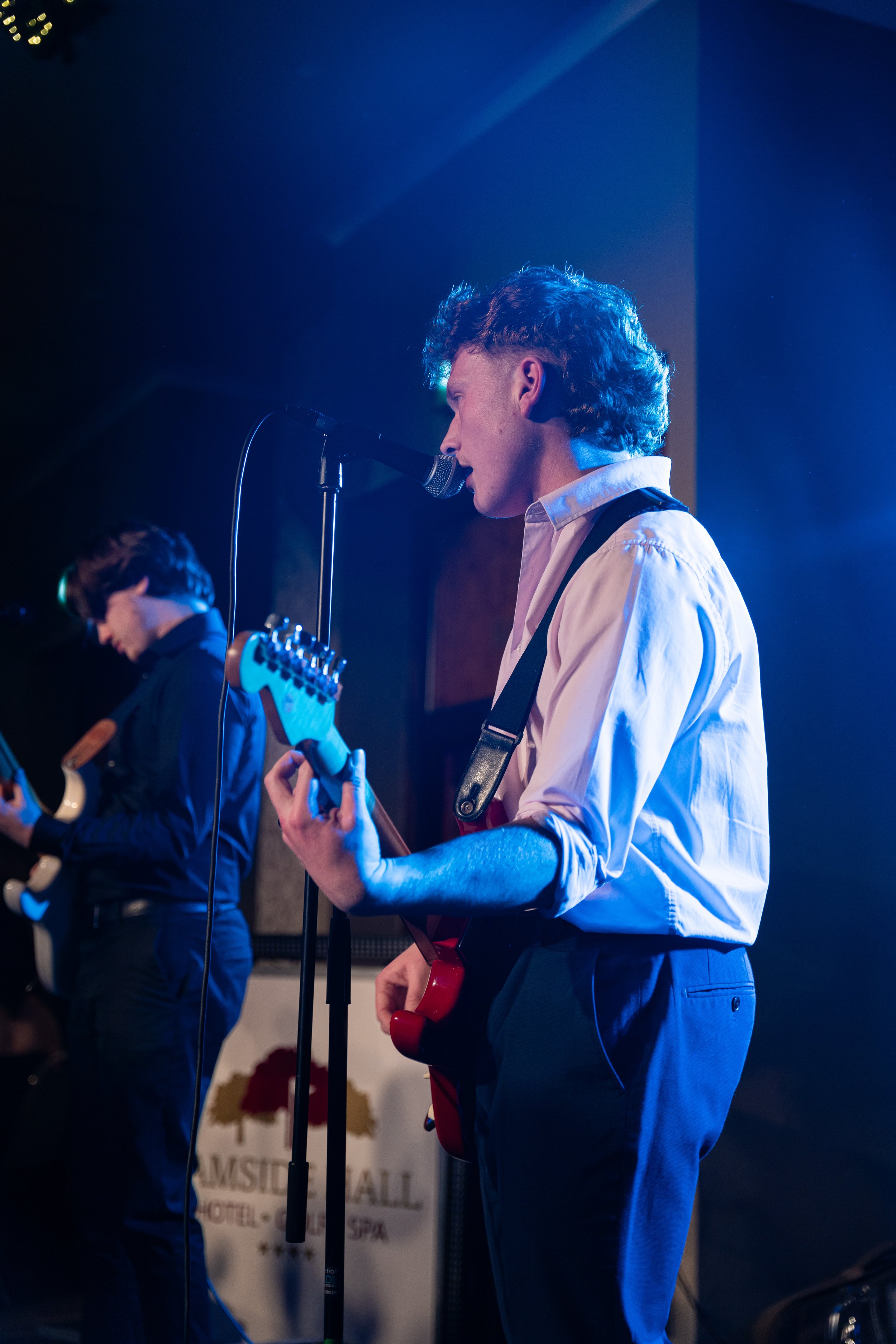 Two musicians performing on stage, with one singing into a microphone and playing an electric guitar, illuminated by blue stage lighting.