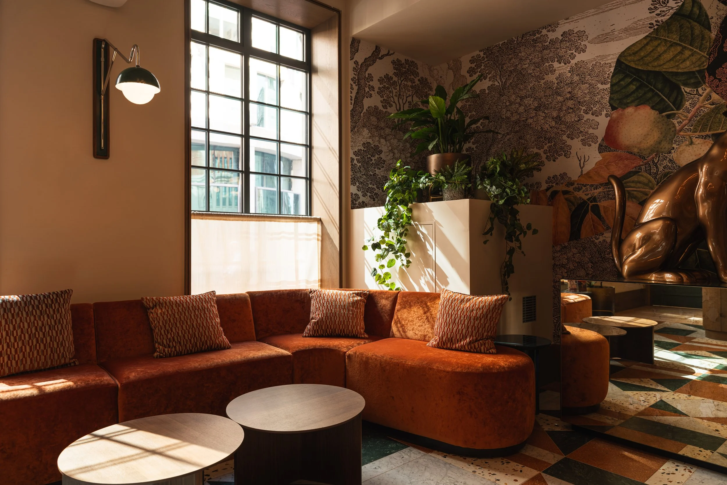 Sunlit interior of a stylish room with a red velvet sectional sofa, patterned throw pillows, a large window with grid panes, potted plants, and a decorative wall with botanical mural.