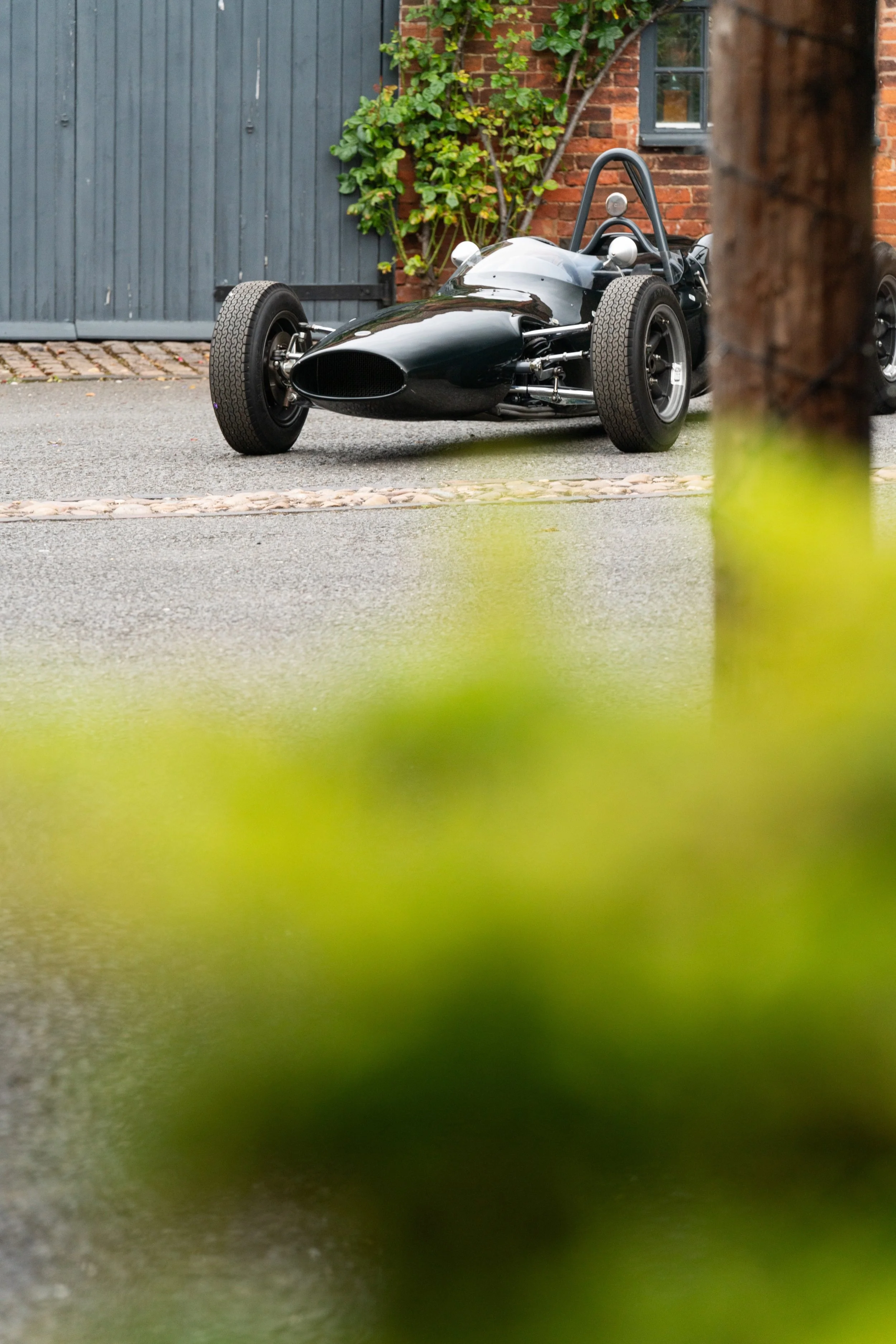 A black vintage race car parked on a paved driveway with brick wall and vine in the background