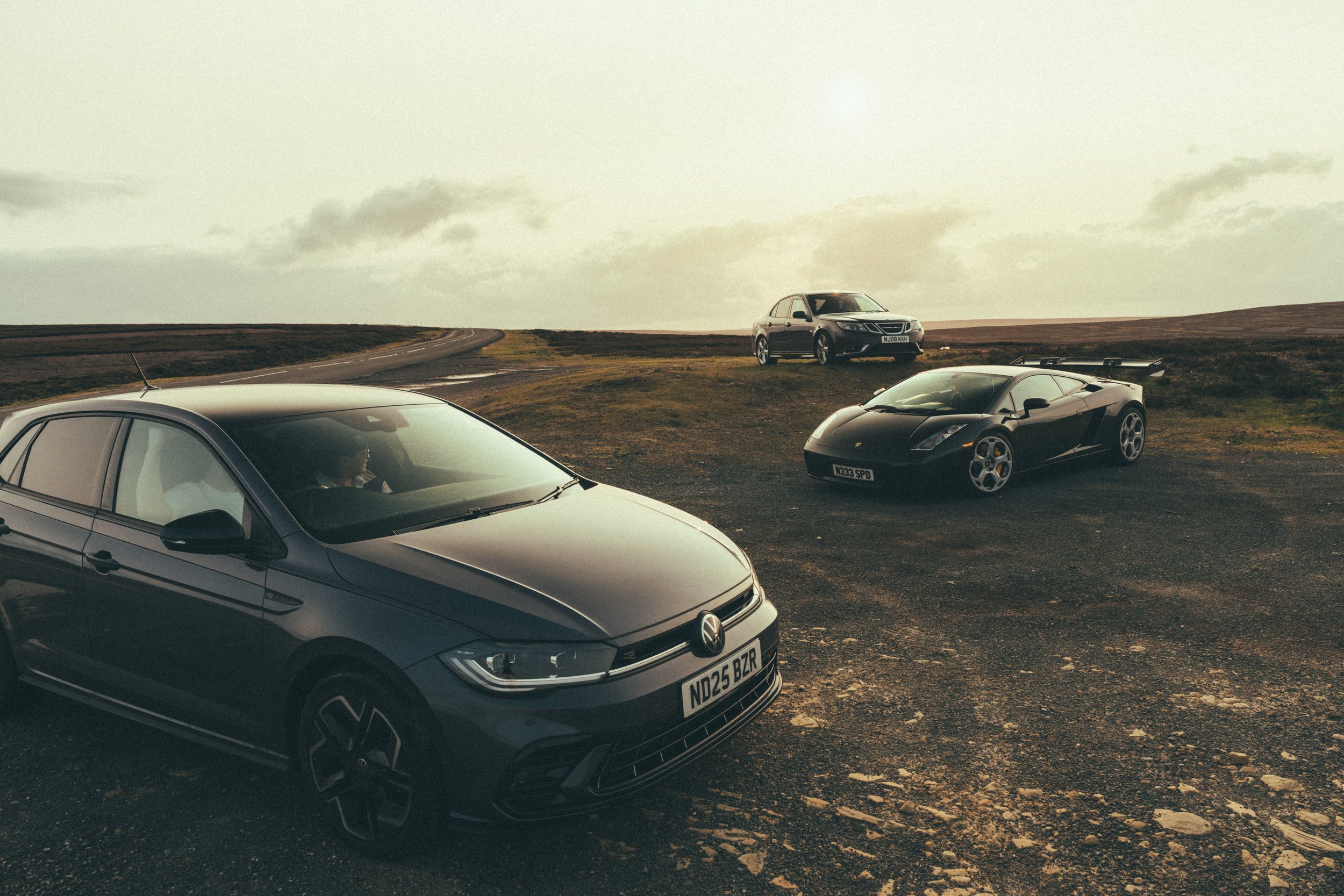 Three cars parked on a countryside landscape with a winding road and cloudy sky. The closest car is a dark Volkswagen sedan, the middle car is a black Lamborghini sports car, and the farthest car is a gray SUV.