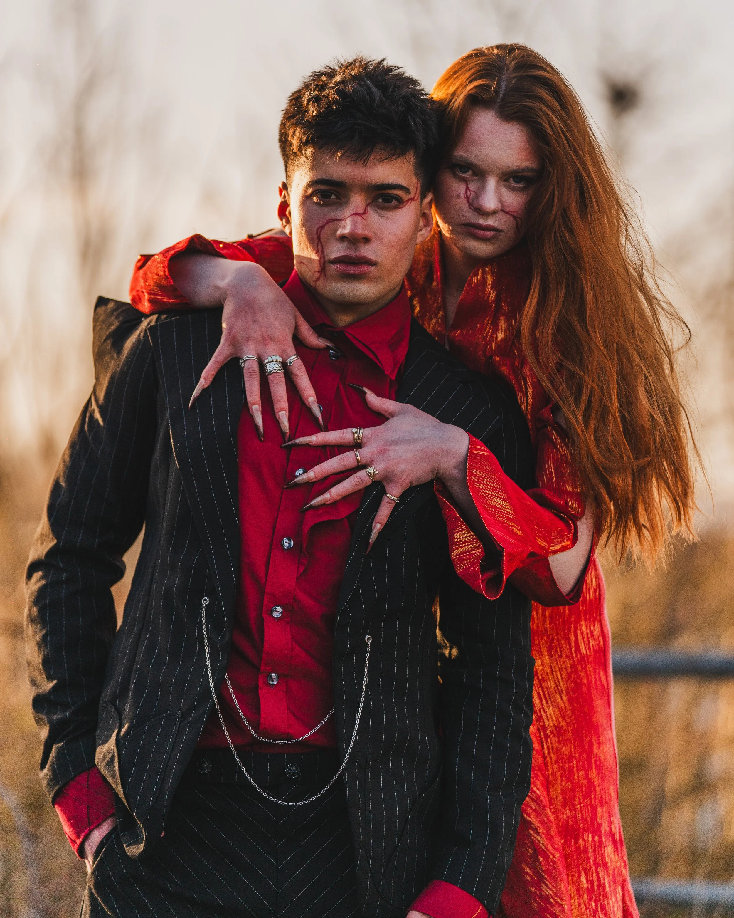 A young man and woman with a dark background stand close together. The woman has long, red hair and wears a shiny red dress, while the man has short, dark hair and wears a black pinstripe suit with a red shirt. They both have scratches or scars on th