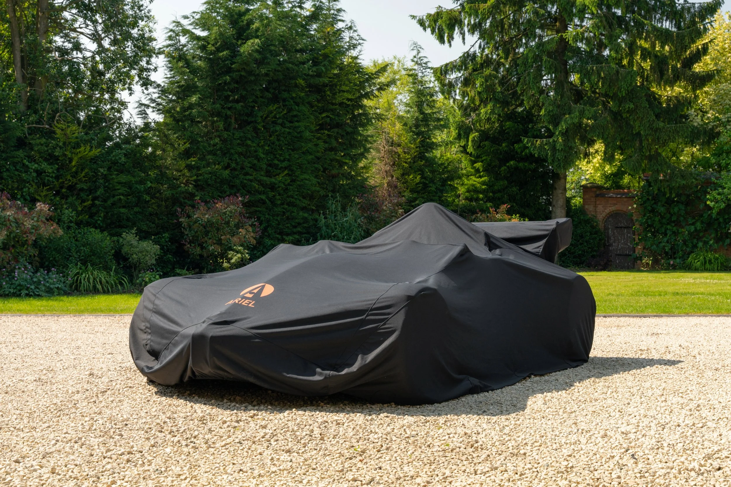 Car covered with black car cover in a garden with lush trees and greenery.