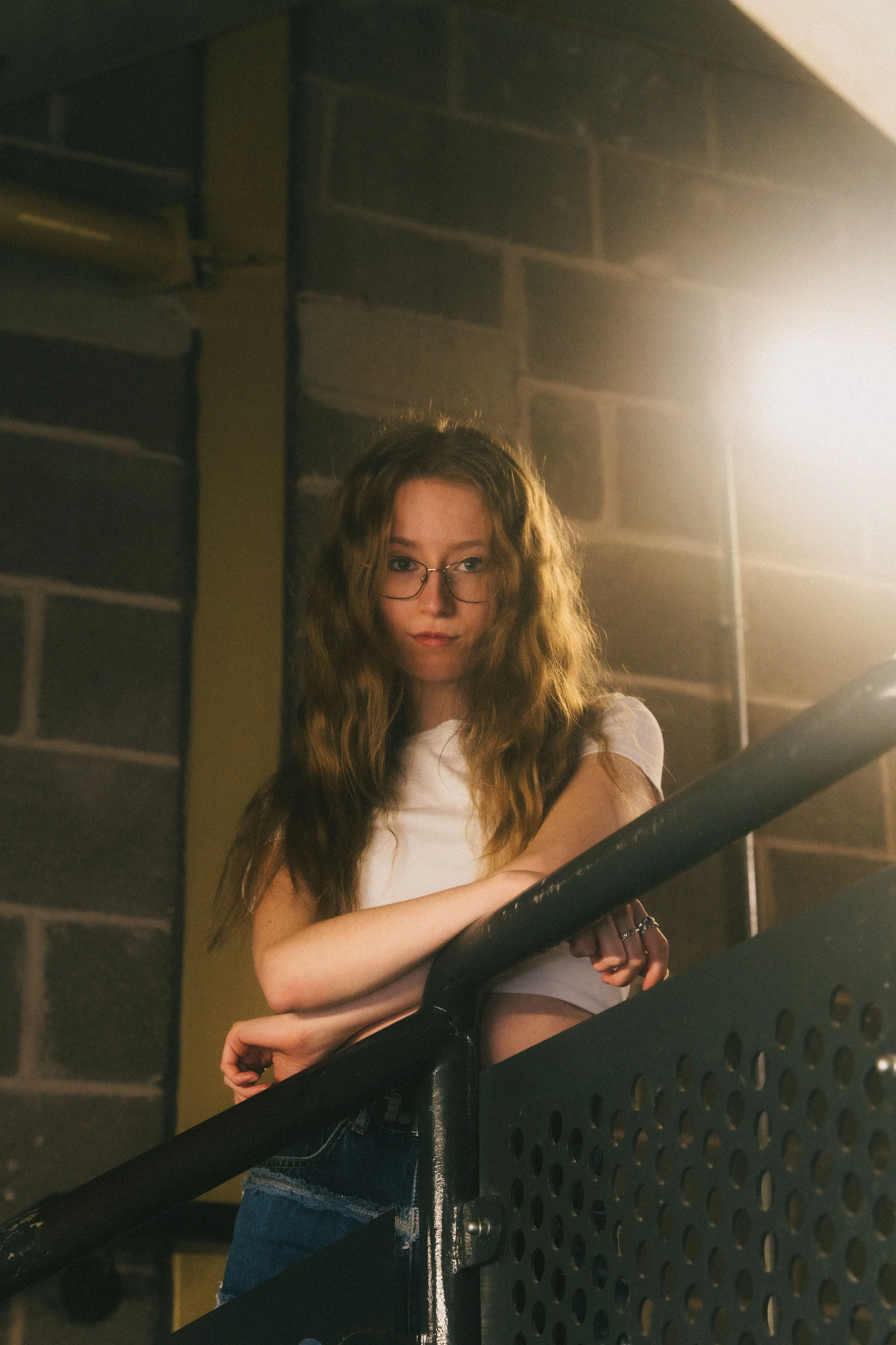 A young woman with long, wavy red hair and glasses, leaning on a black railing, indoors near a brick wall and a bright light.