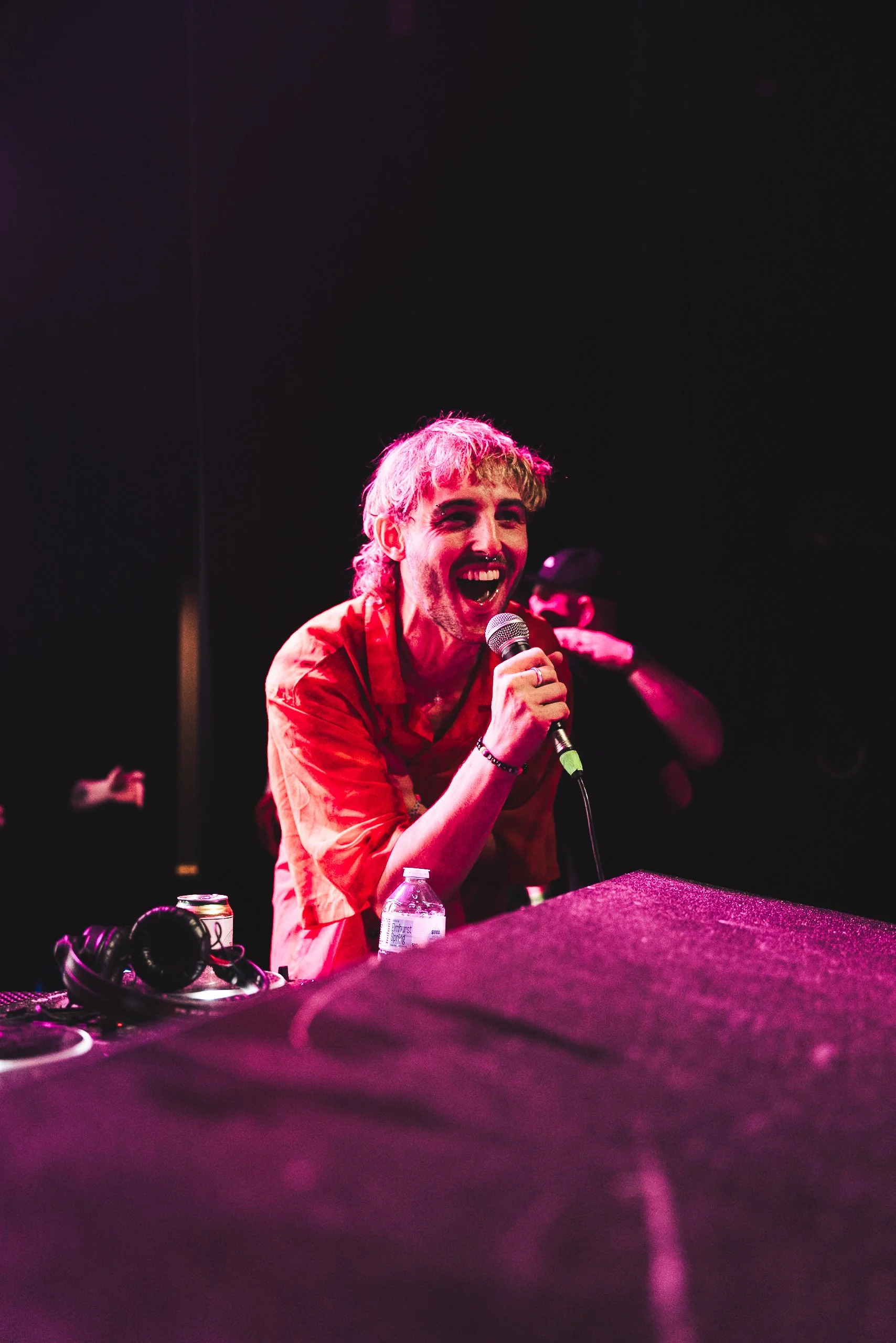 A person with blonde hair singing into a microphone on stage, smiling, with DJ equipment, a water bottle, and a can nearby.