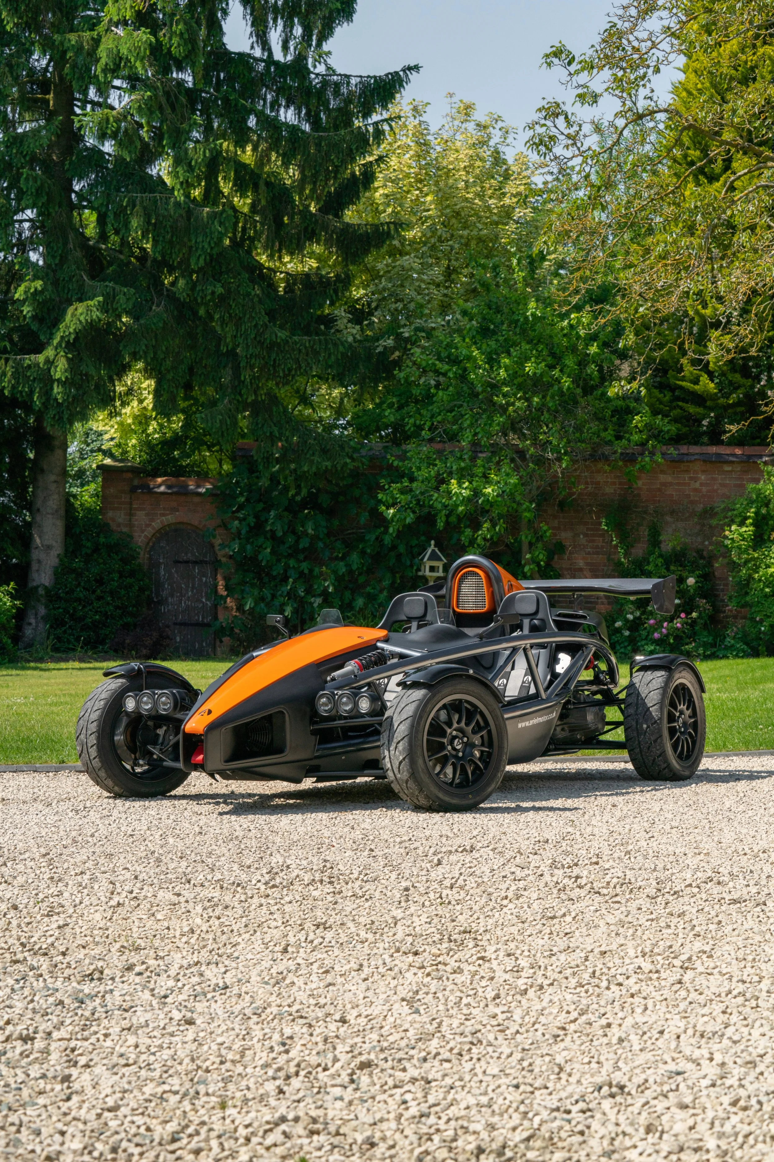 A black and orange open-wheel race car parked on a gravel driveway in a lush green garden with trees and a brick wall in the background.