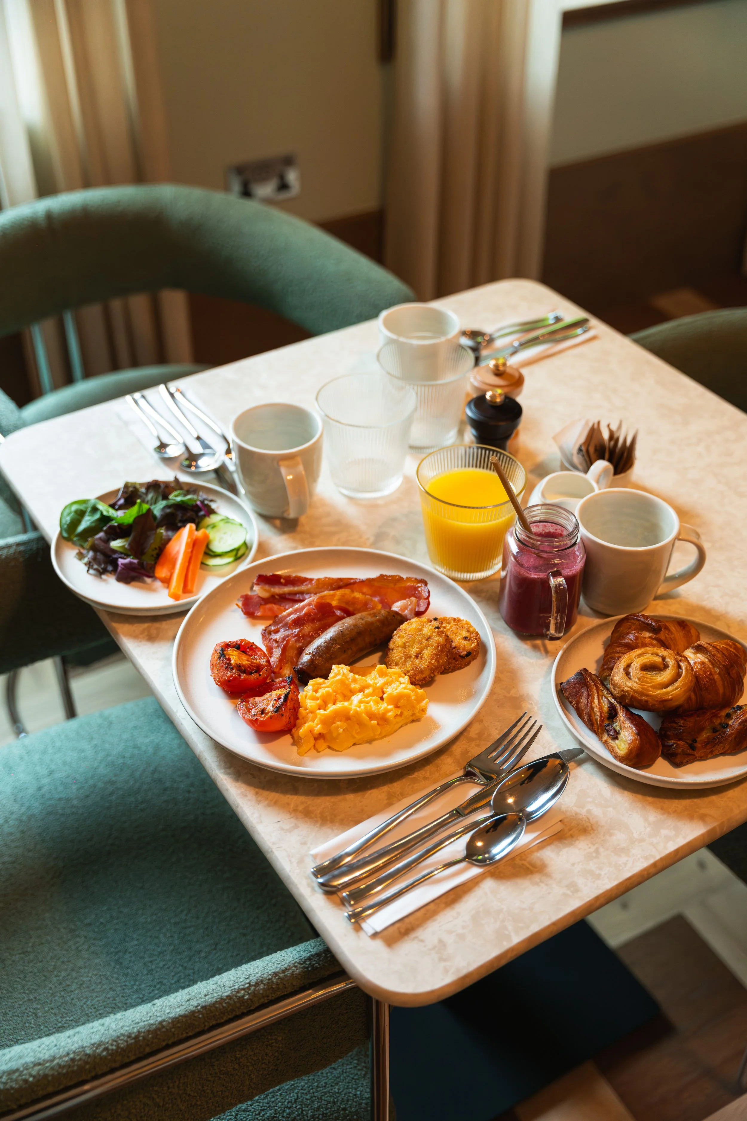 A breakfast table with scrambled eggs, bacon, sausage, tomatoes, a salad, pastries, orange juice, coffee, and various condiments, set with forks, knives, and napkins.