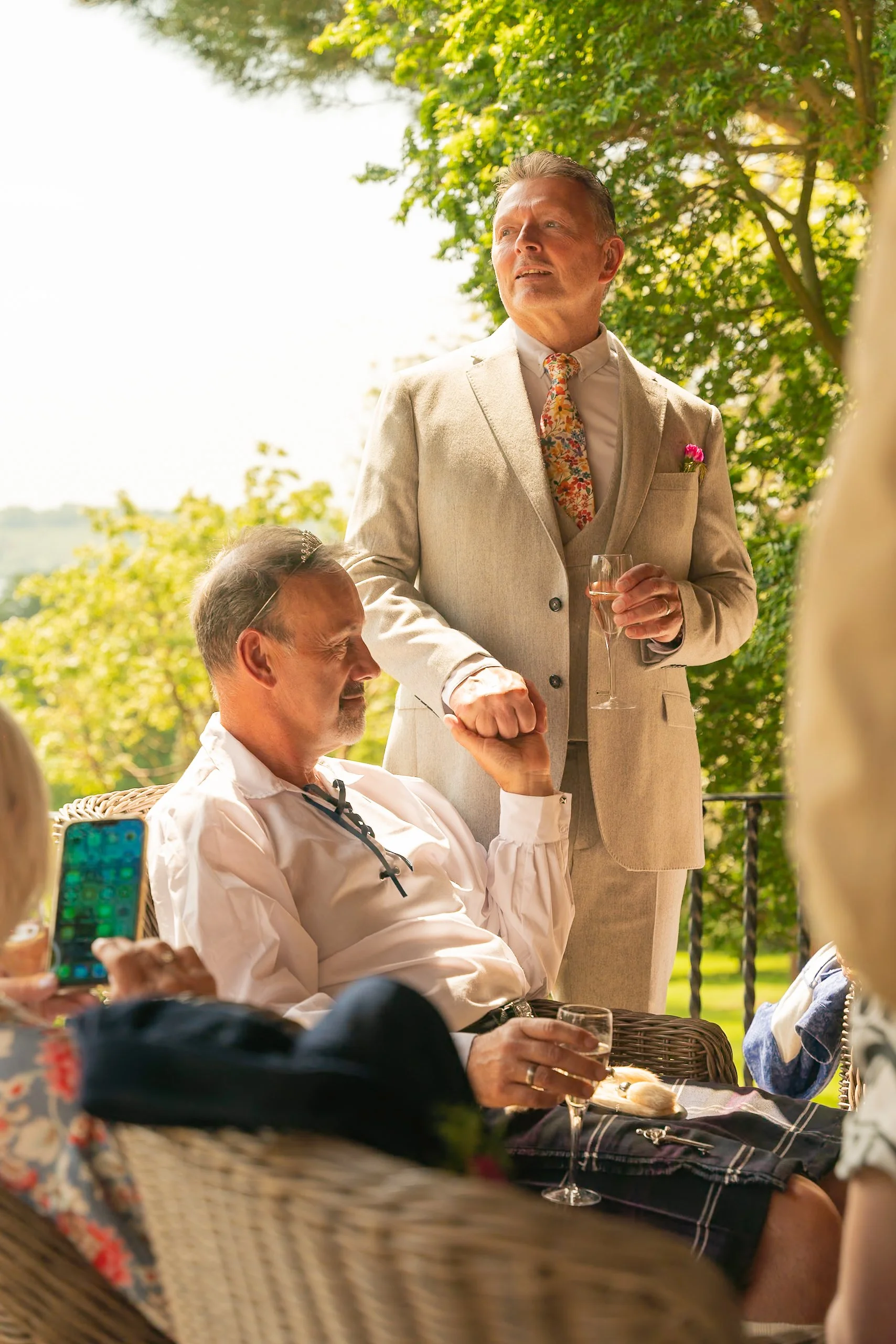Man in traditional Scottish attire sitting on a wicker chair, holding a glass of wine, and taking a selfie with a smartphone. Another man in a light-colored suit stands nearby, holding a glass of champagne, during an outdoor gathering on a sunny day 