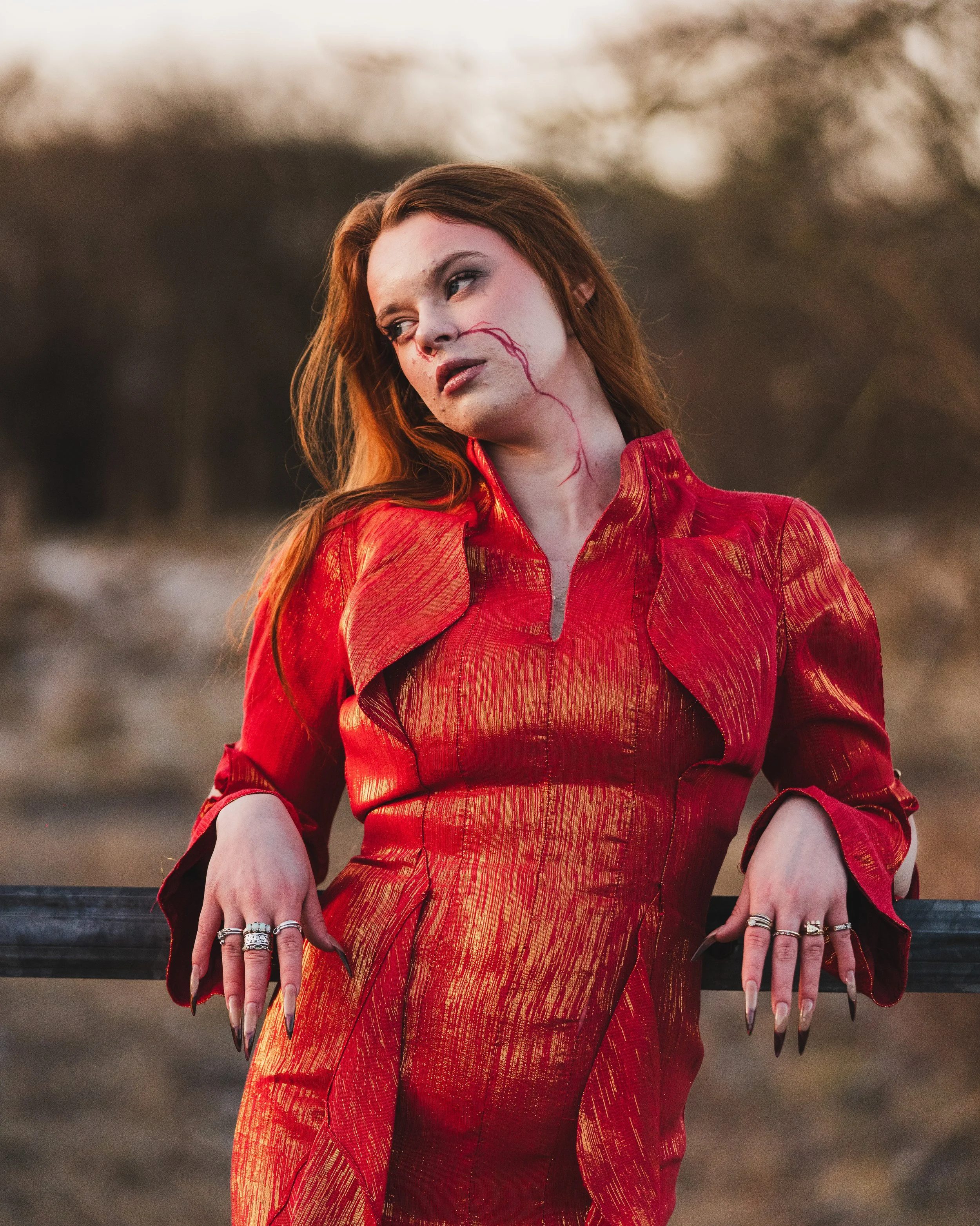 A woman with long red hair and pale skin wearing a shiny red dress with textured gold accents, leaning on a black metal fence outdoors. She has dark makeup, long black nails, and rings on her fingers. She appears to be lost in thought, with a somber 