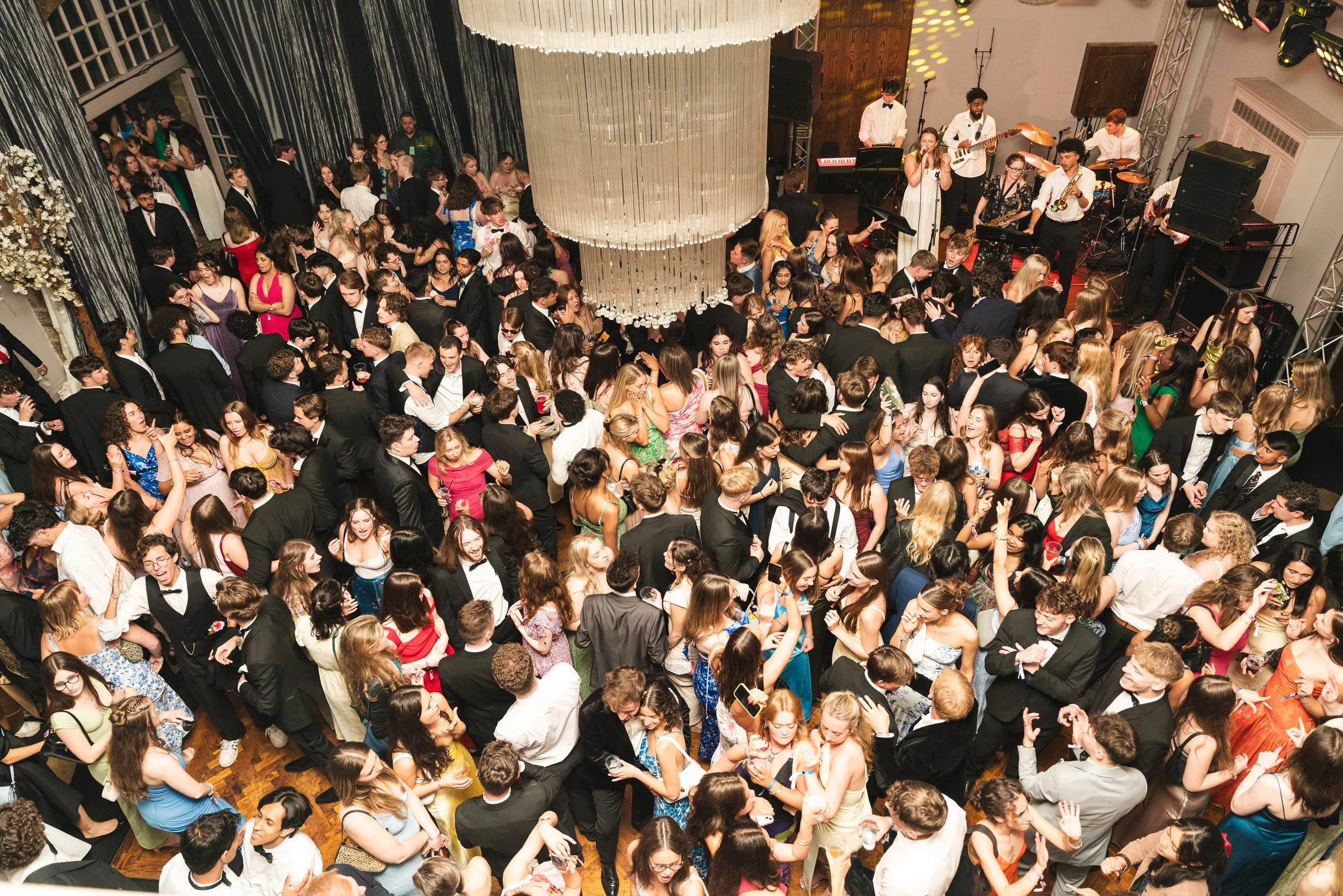 Crowd of young adults dancing and socializing at a formal event with a live band performing on stage.