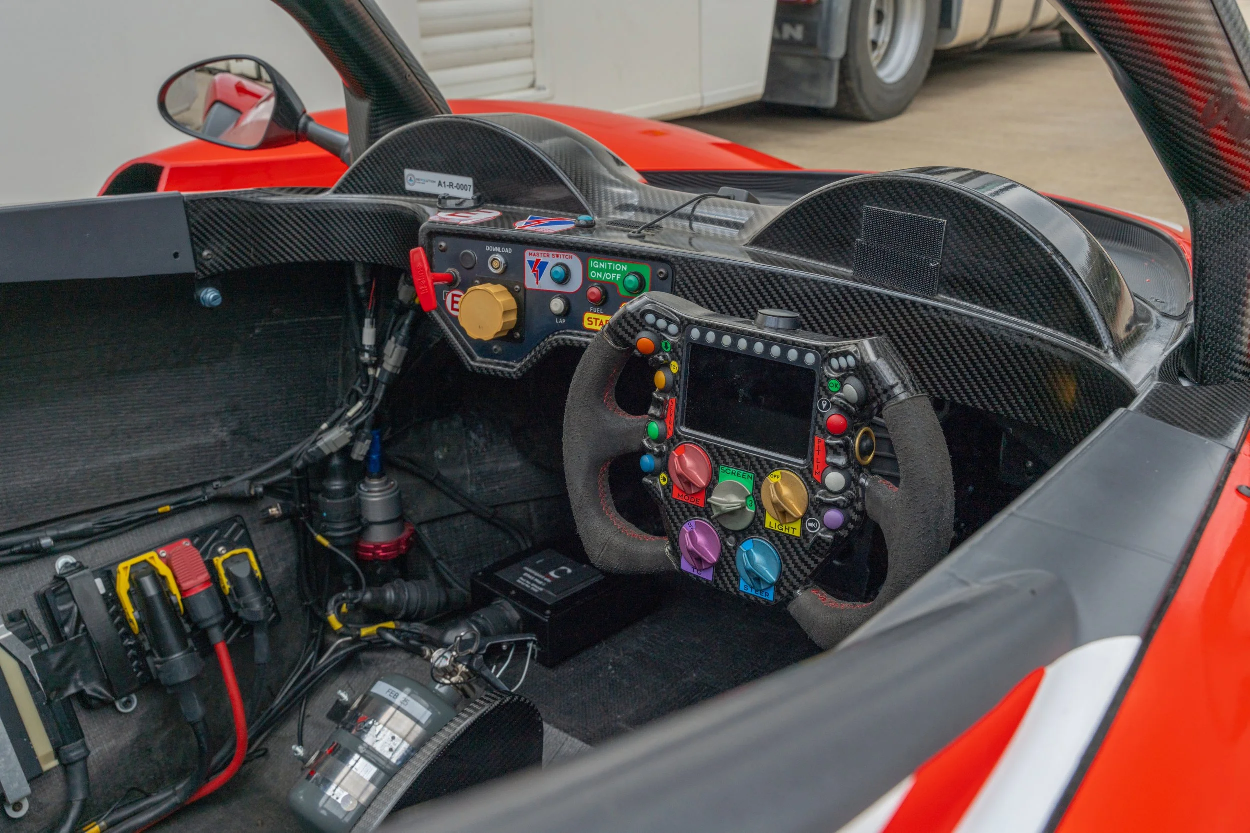 Interior of a race car cockpit with a complex dashboard and steering wheel featuring various colorful buttons and switches, made of carbon fiber with a red exterior visible.