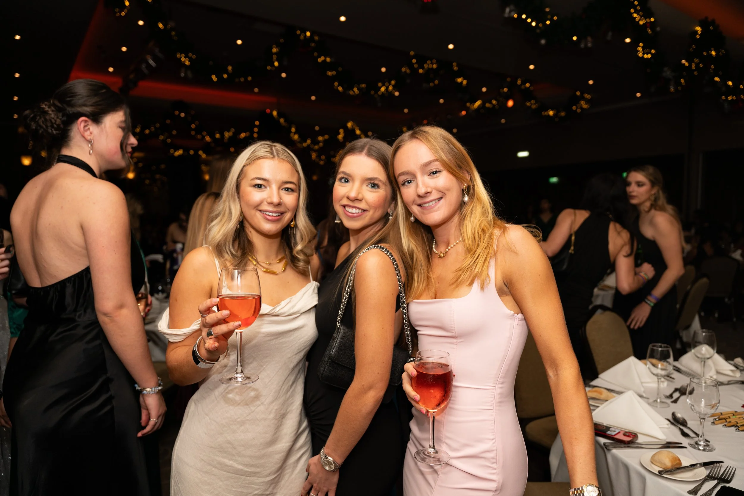 Four young women in elegant dresses at a social event, smiling at the camera while holding glasses of pink-colored wine, with tables set for a meal in the background and festive decorations hanging from the ceiling.