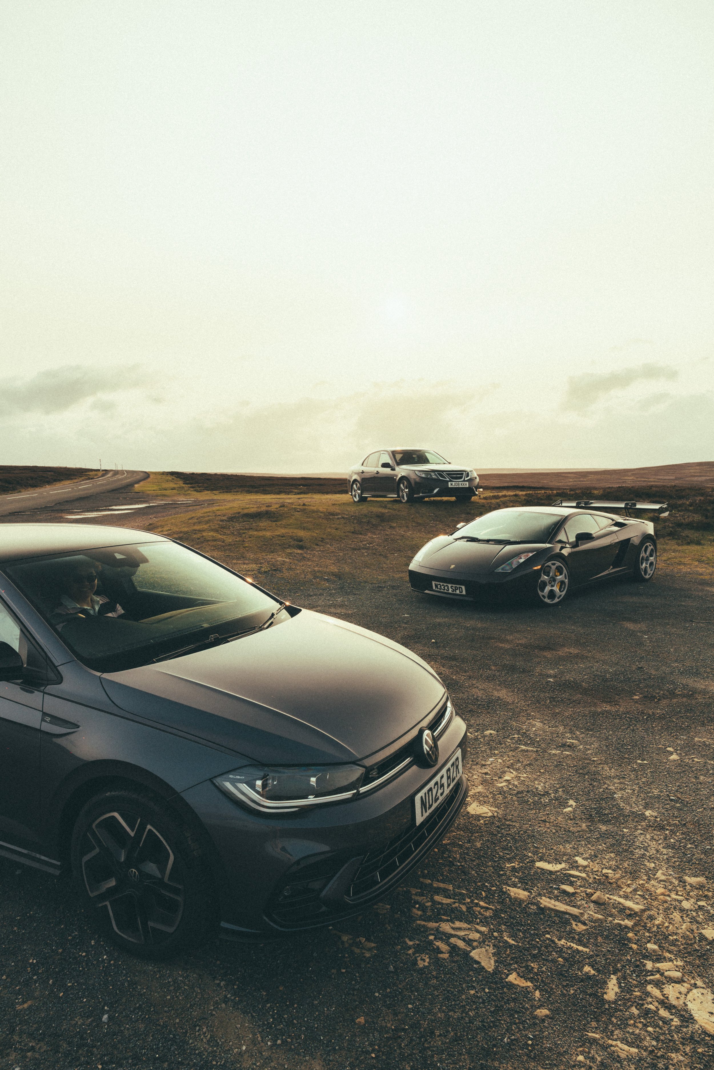 Three parked cars on a barren landscape with a cloudy sky, including a Volkswagen in the foreground, a black sports car with a spoiler, and a silver sedan in the background.