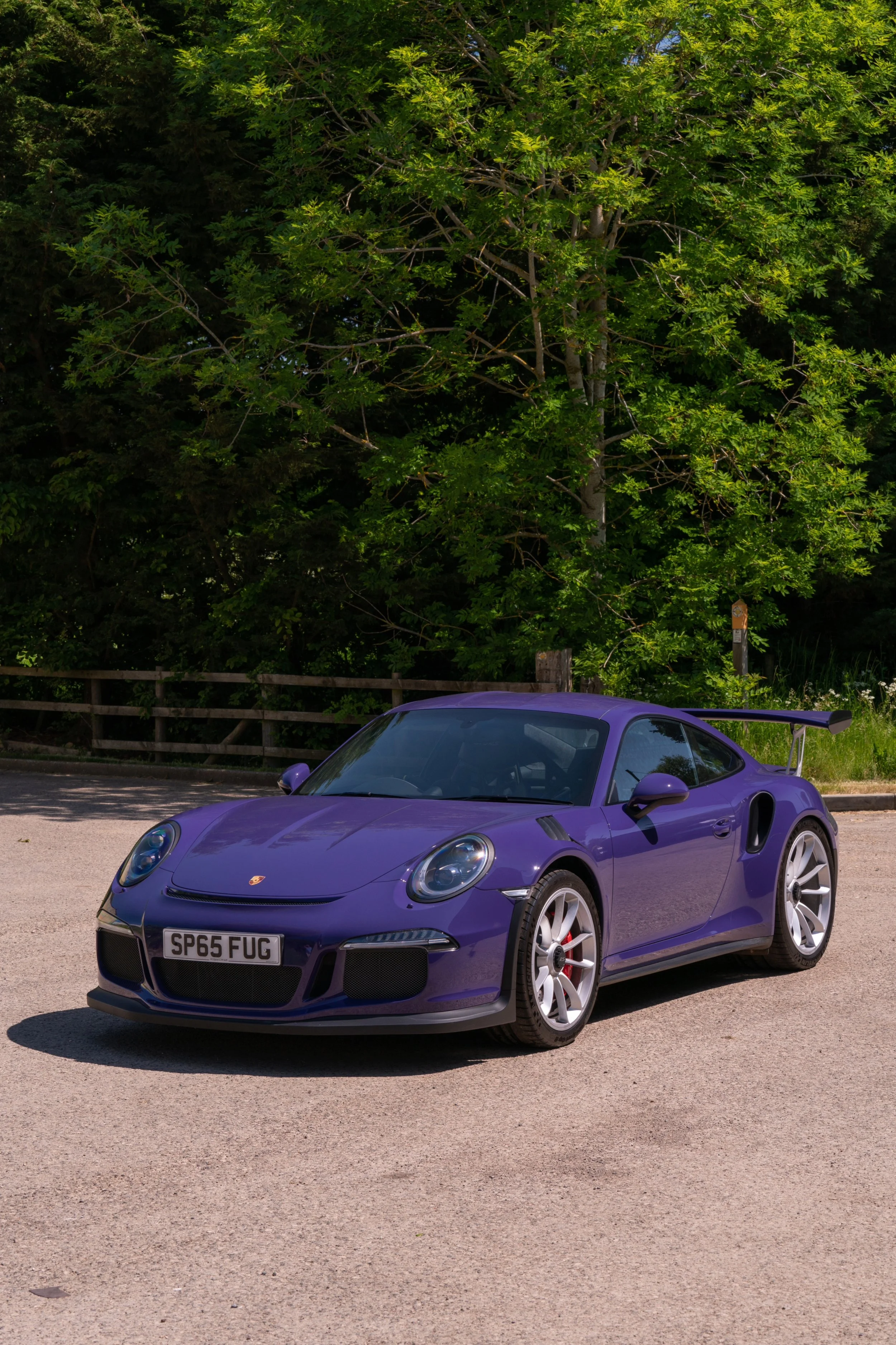 A purple Porsche sports car parked on a gravel road near a green tree, under a blue sky.