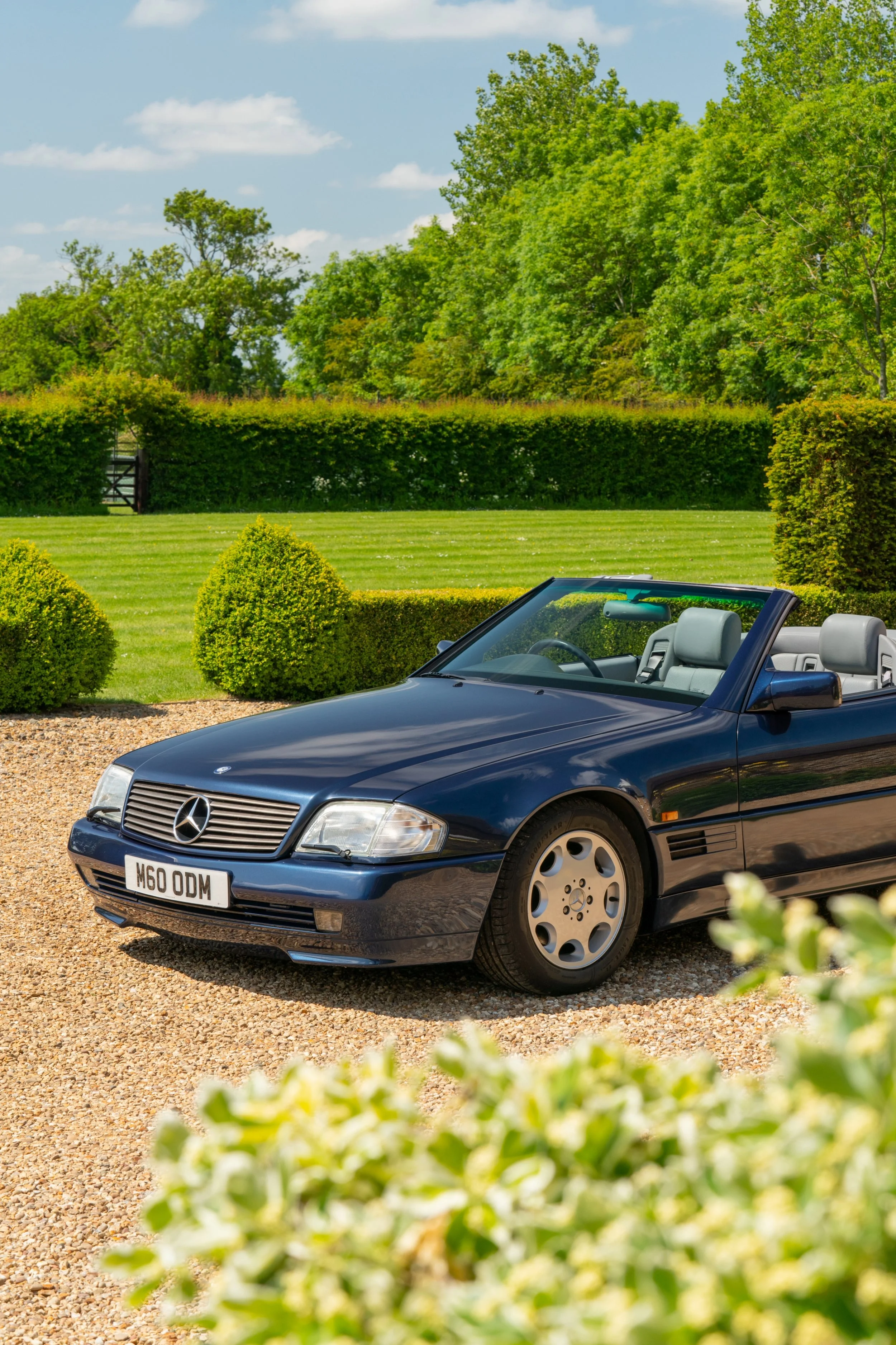 A classic blue Mercedes-Benz convertible parked on a gravel driveway with green manicured lawn, bushes, and trees in the background under a partly cloudy sky.