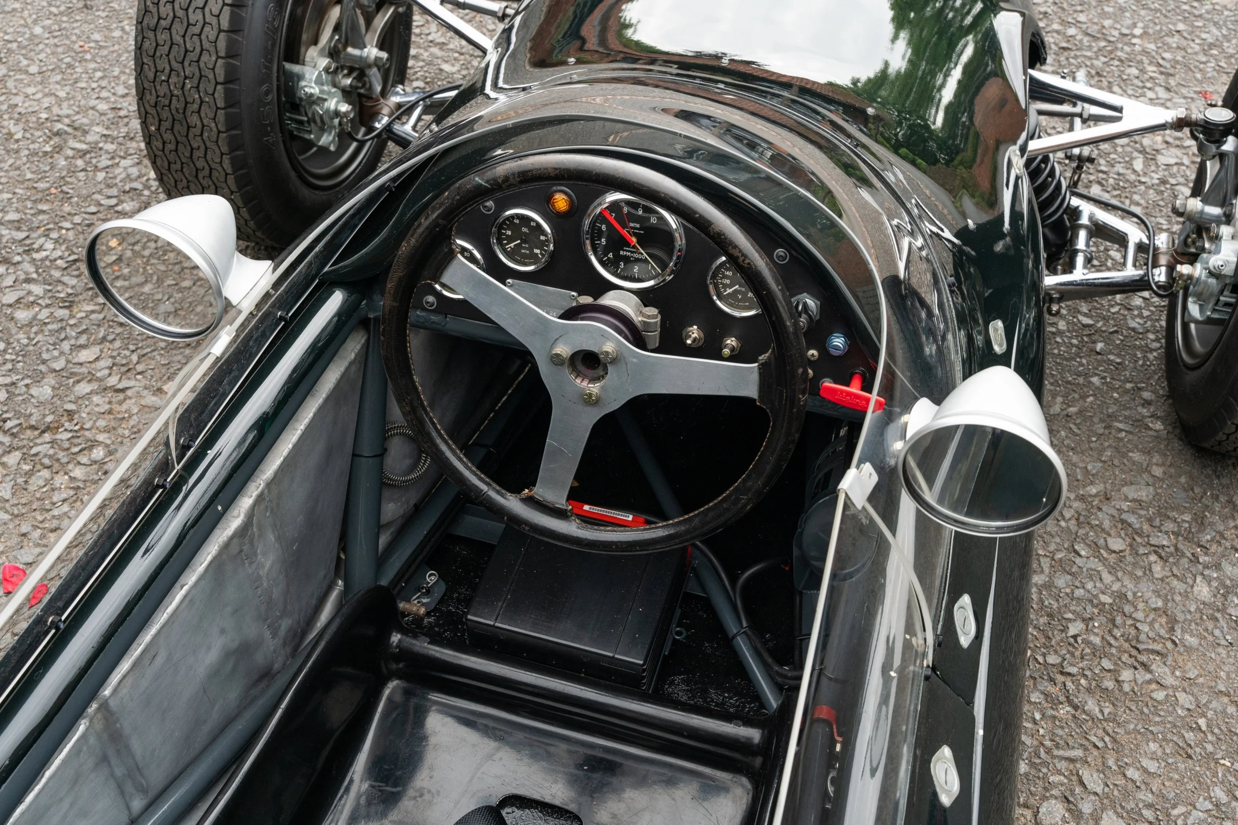 Interior view of a vintage race car showing a black steering wheel, analog gauges including a tachometer, and a small windshield with side mirrors, on a gravel surface.